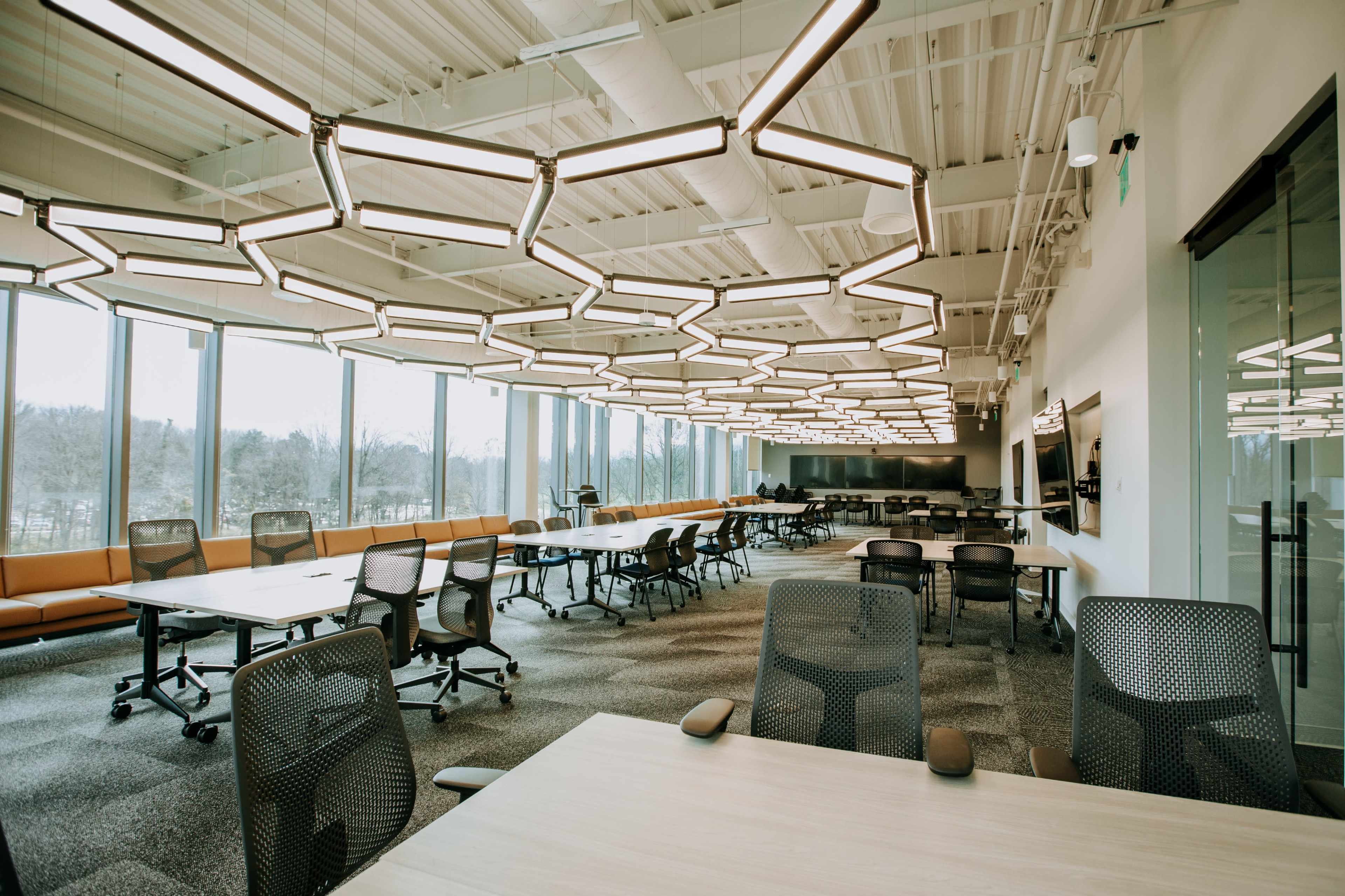 The image shows a modern office space with multiple tables and chairs, large windows, and unique overhead lighting.