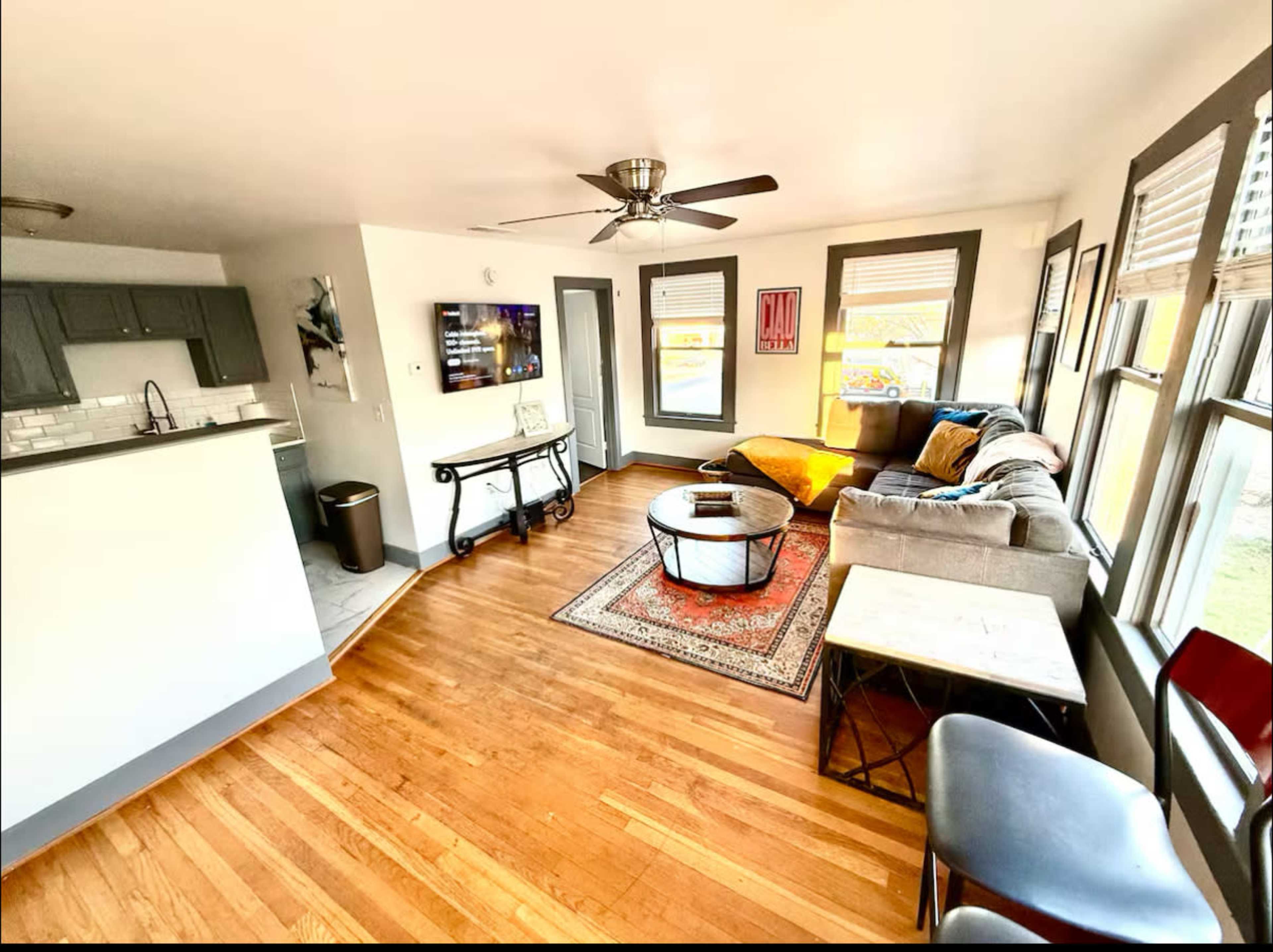 The image shows a bright living room featuring a gray sectional couch, a round coffee table, a TV mounted on the wall, and a small kitchenette at one end.