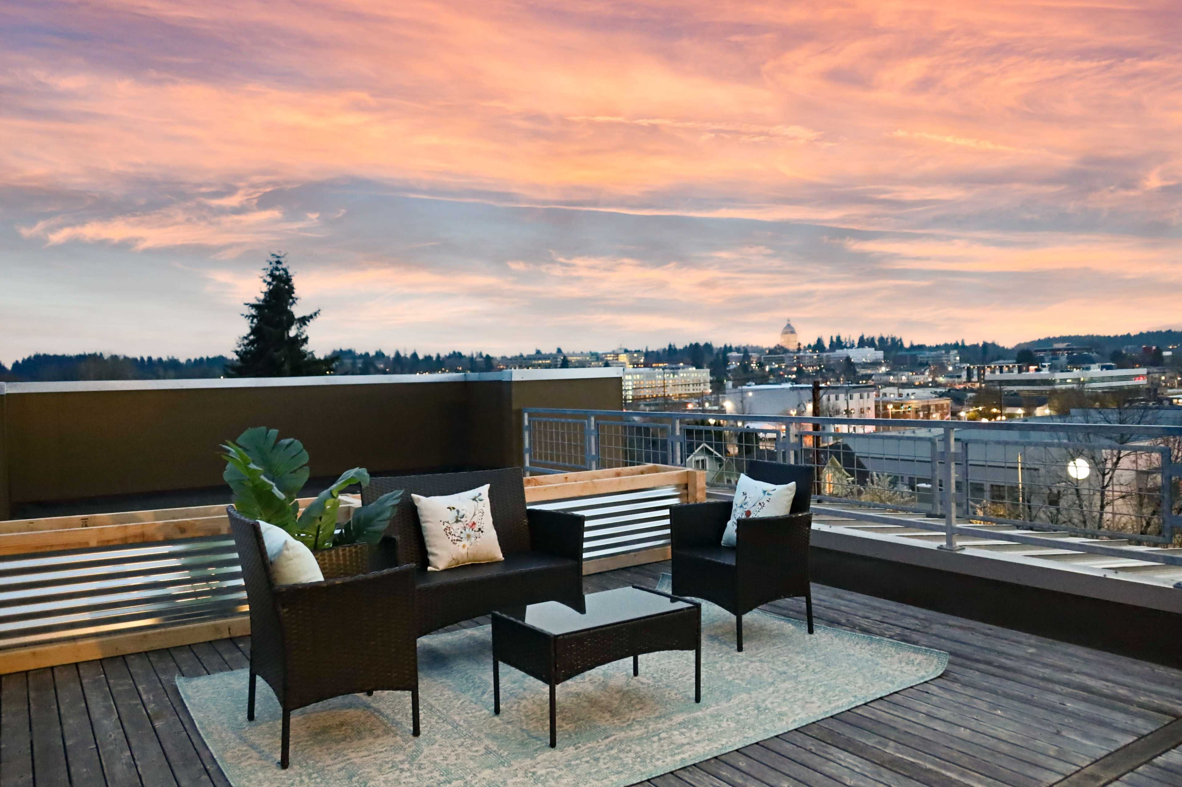 A rooftop terrace features a set of outdoor furniture, including two chairs and a coffee table, overlooking a cityscape at sunset.