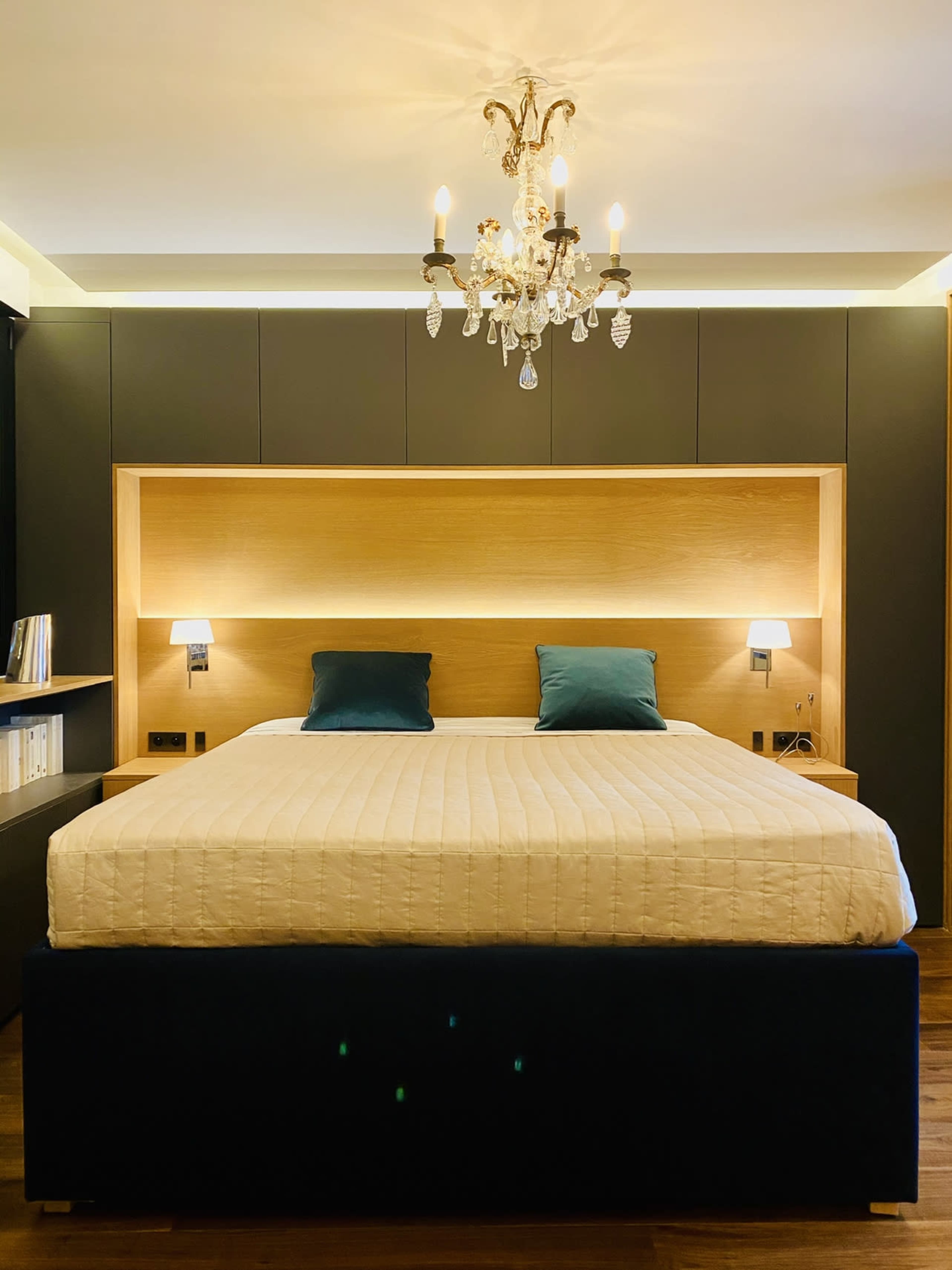 A modern bedroom features a large bed with blue accent pillows, a wooden headboard, and a chandelier above.