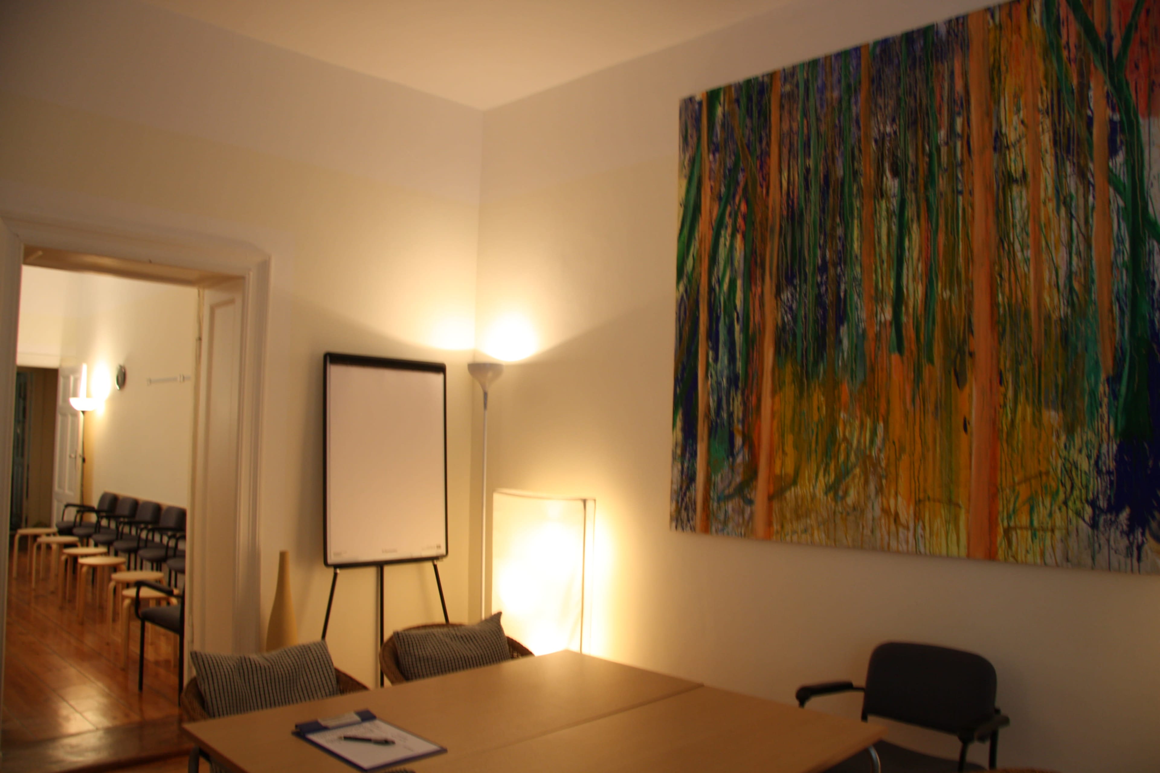 The image shows a well-lit room featuring a table and chairs, with an easel board and a large colorful painting on the wall.