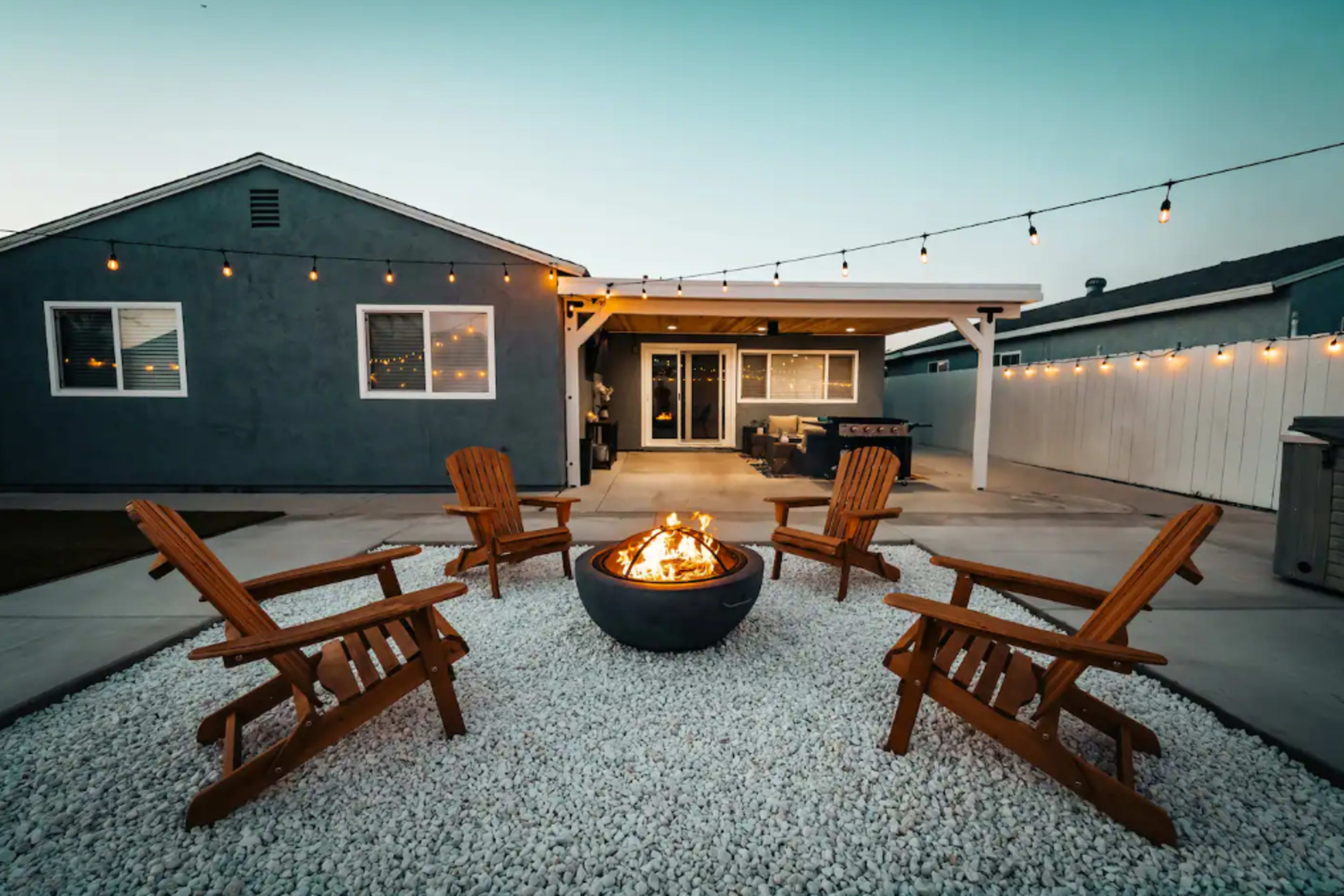 A backyard featuring a fire pit surrounded by four wooden chairs, with string lights illuminating the area.