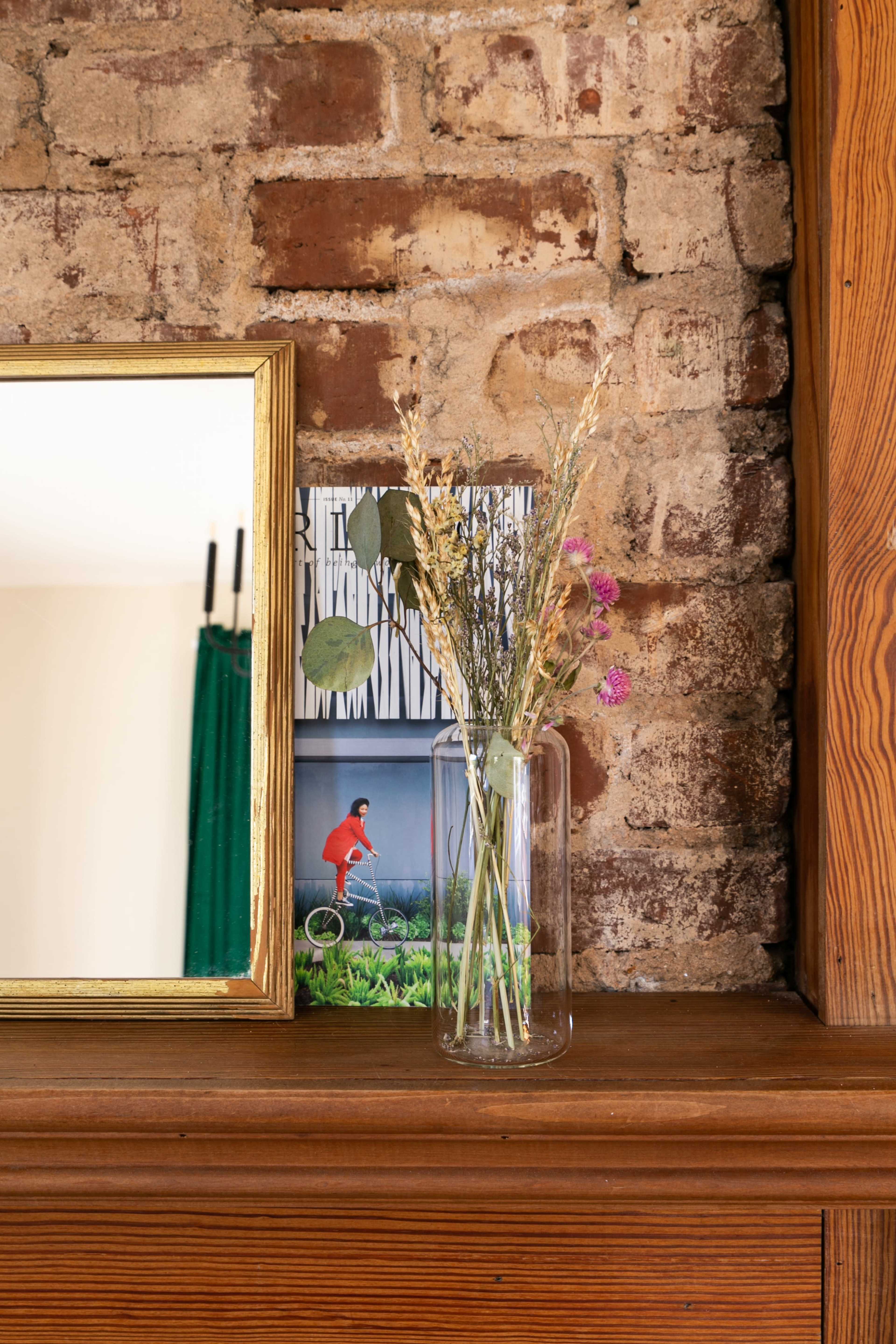 A glass vase with dried flowers stands on a wooden shelf next to a mirror and a framed image against a brick wall.