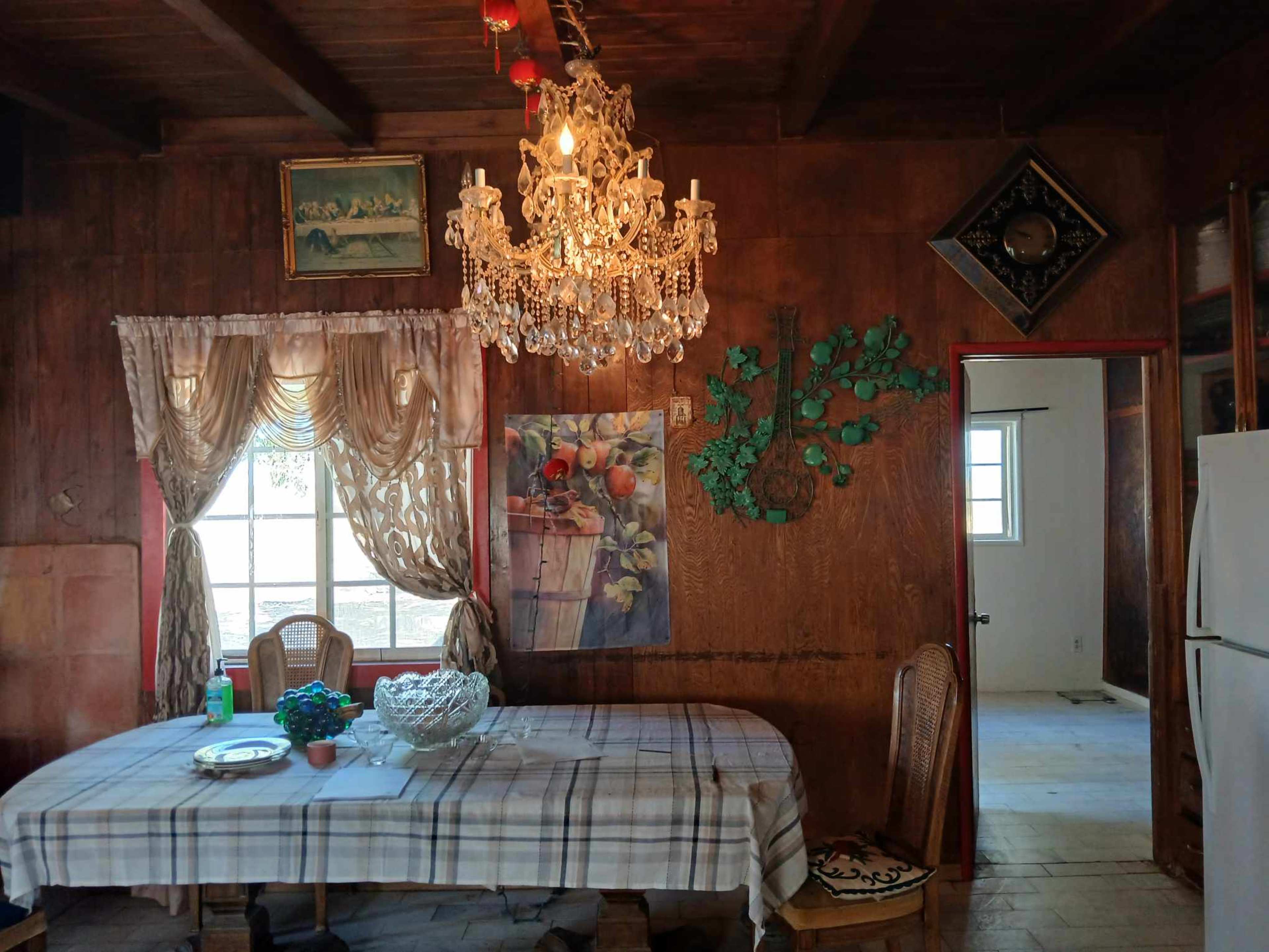 The interior of a rustic dining room features a wooden table covered with a plaid tablecloth, a chandelier hanging from the ceiling, and decorative pieces on the walls.