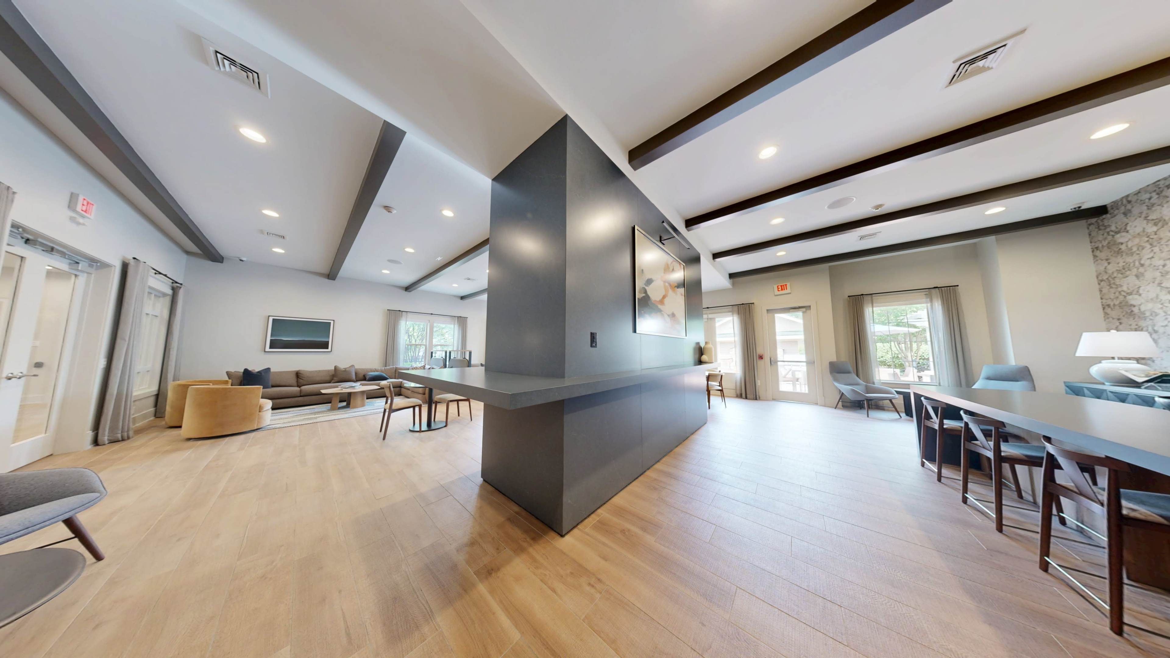 The image shows a well-lit, modern communal area featuring a seating arrangement on one side and a dining table on the other, with large windows and wooden flooring.