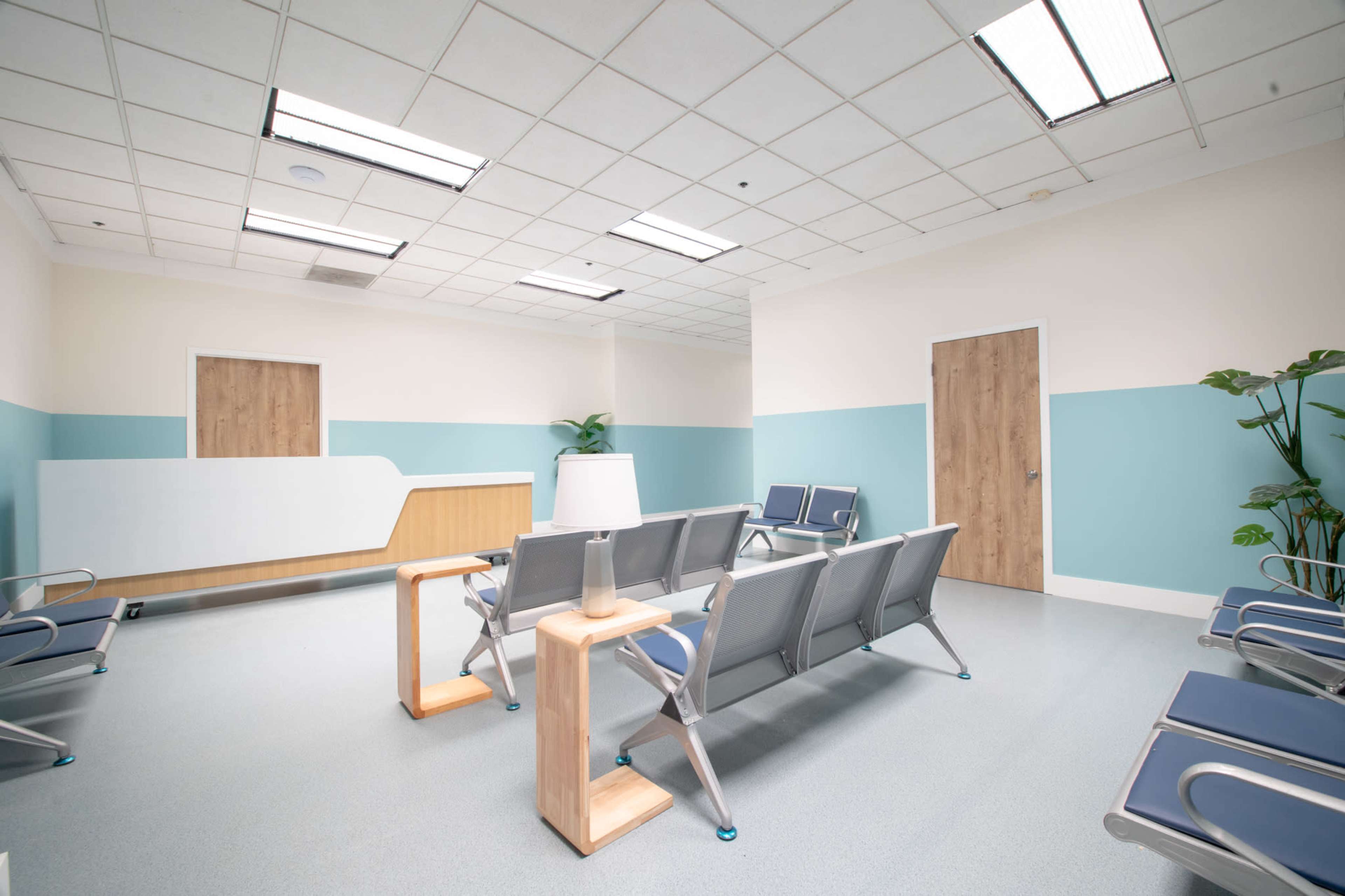 The image shows a clean and modern waiting room with rows of seating, a reception area, and decorative plants.
