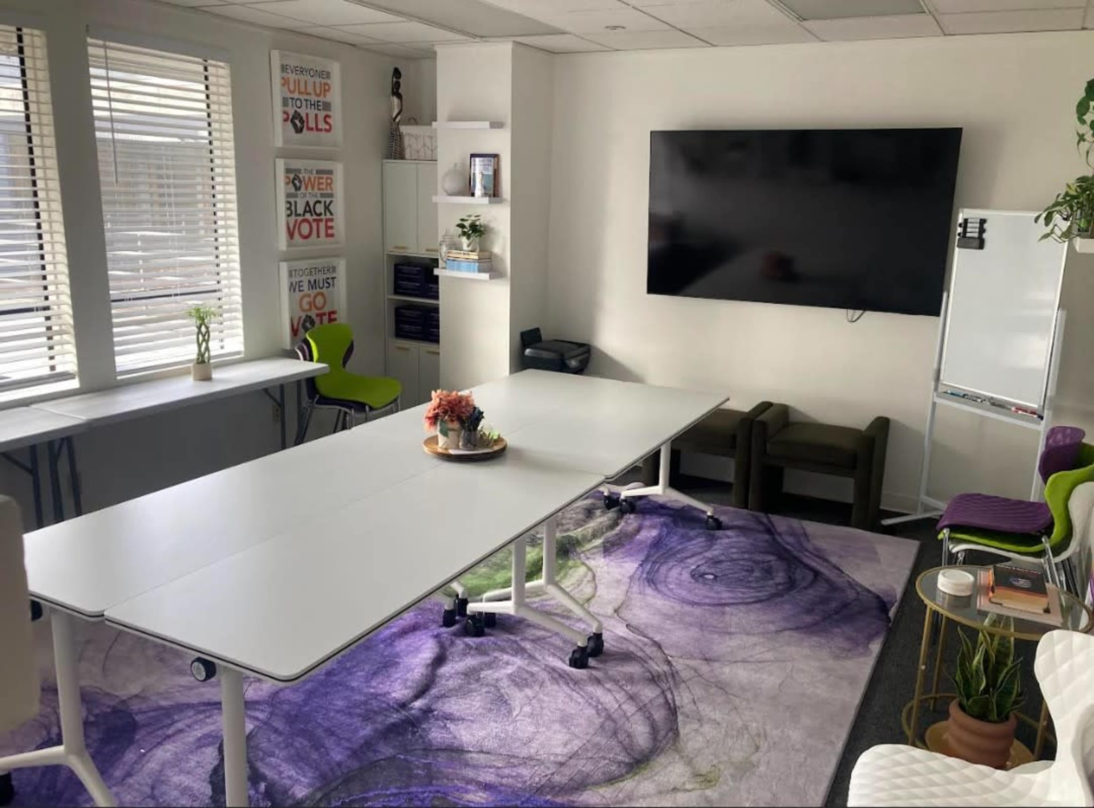 The image shows a bright, modern meeting room featuring a large table, chairs, a wall-mounted television, and a whiteboard, with decorative plants and a purple rug on the floor.