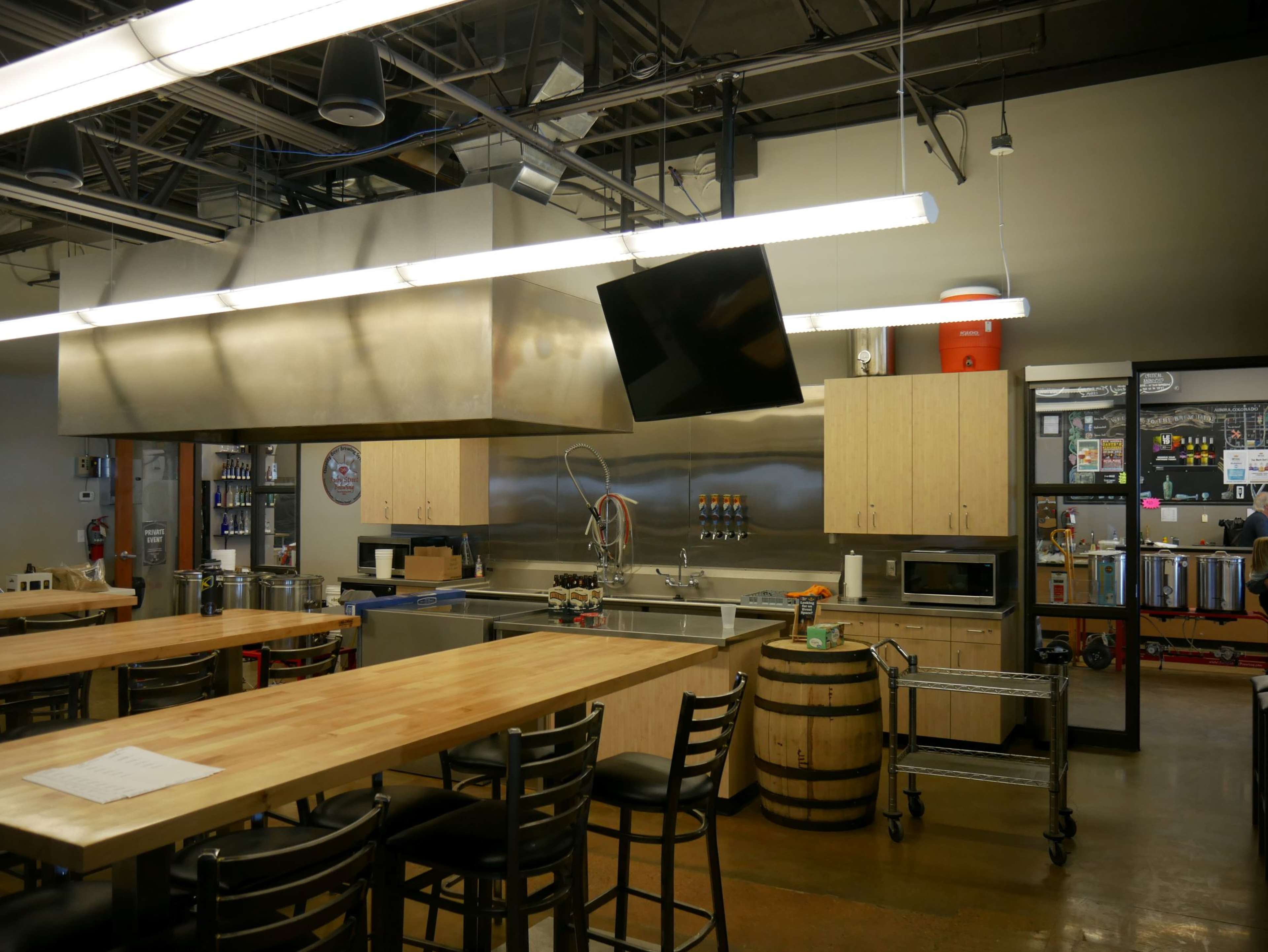 A modern kitchen space with a large central wooden table, stainless steel appliances, and a barrel used as a table.