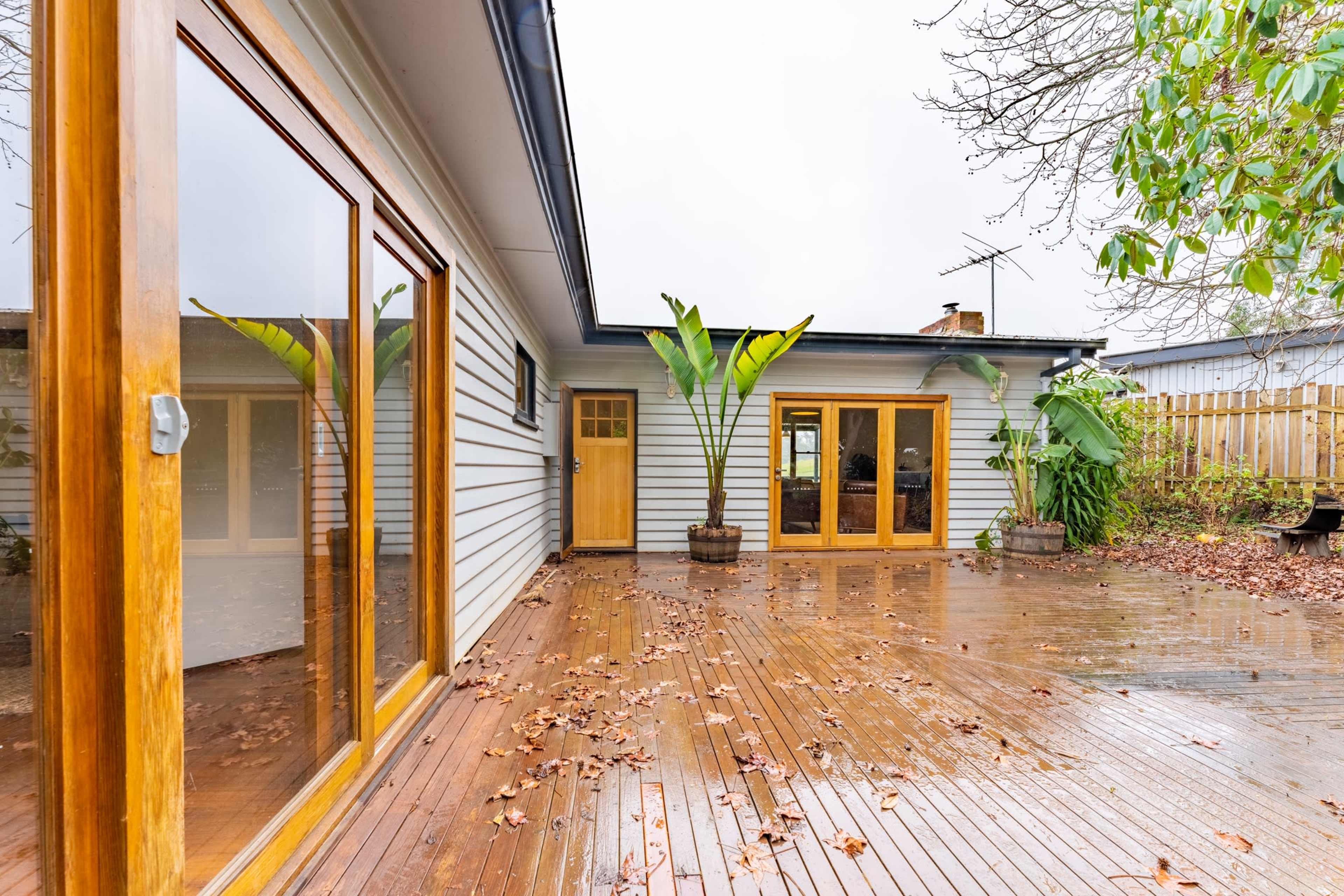 A wooden deck outside a house with large potted plants and some fallen leaves on the wet surface.