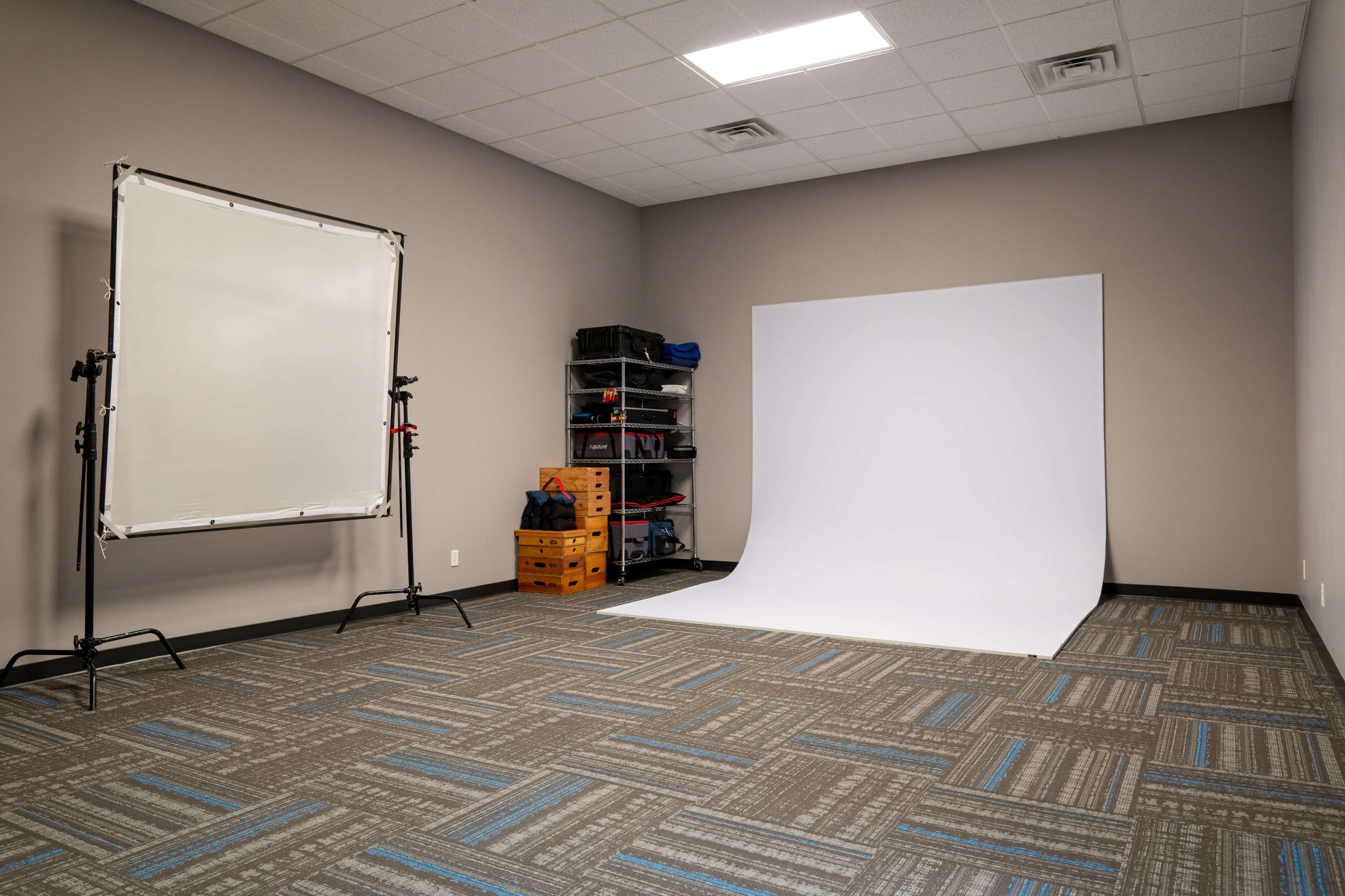 The image shows a photography studio with a white backdrop, a light-diffusing screen on a stand, and storage units filled with equipment.
