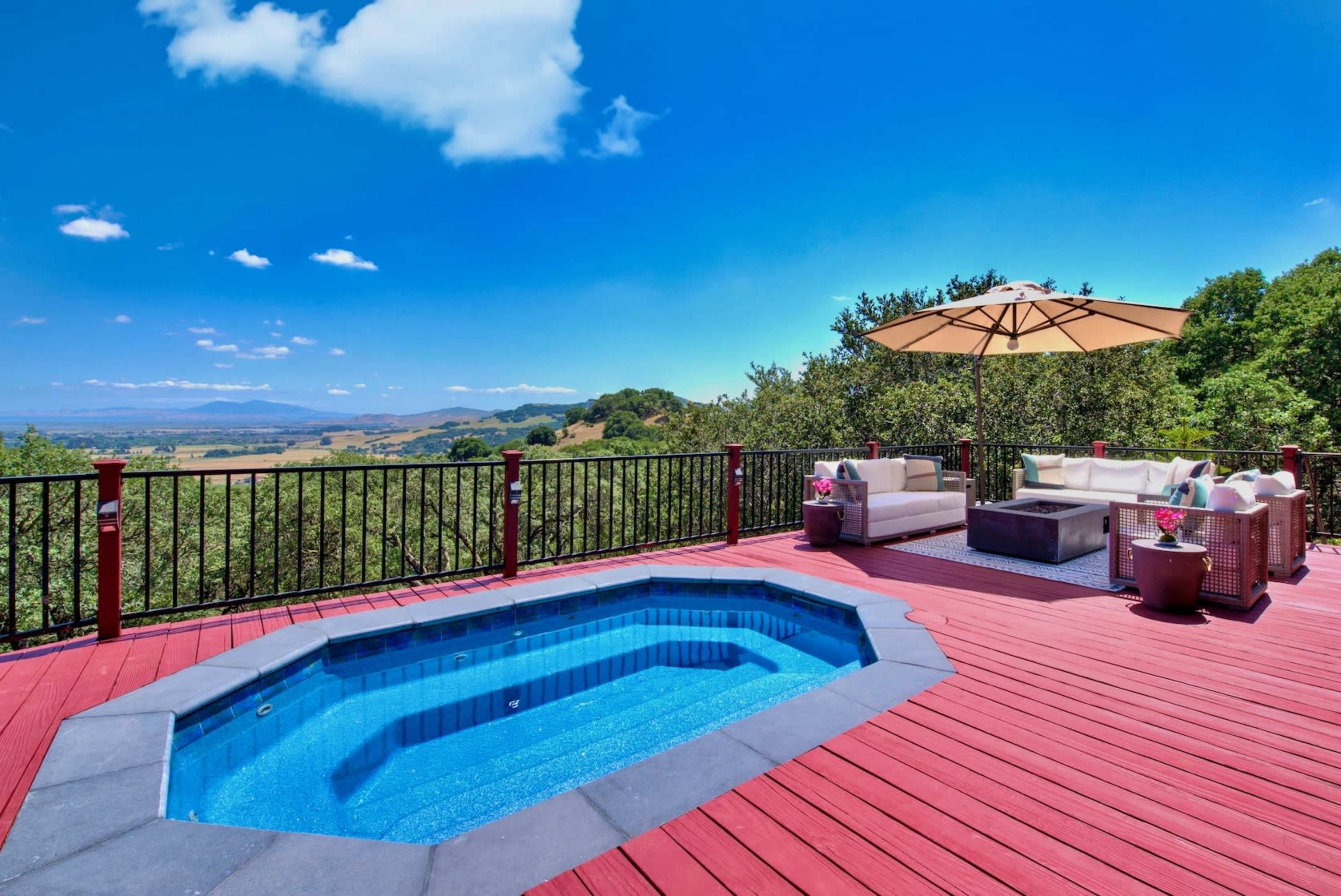 A deck with a hot tub, seating area, and an umbrella overlooks a valley under a clear blue sky.