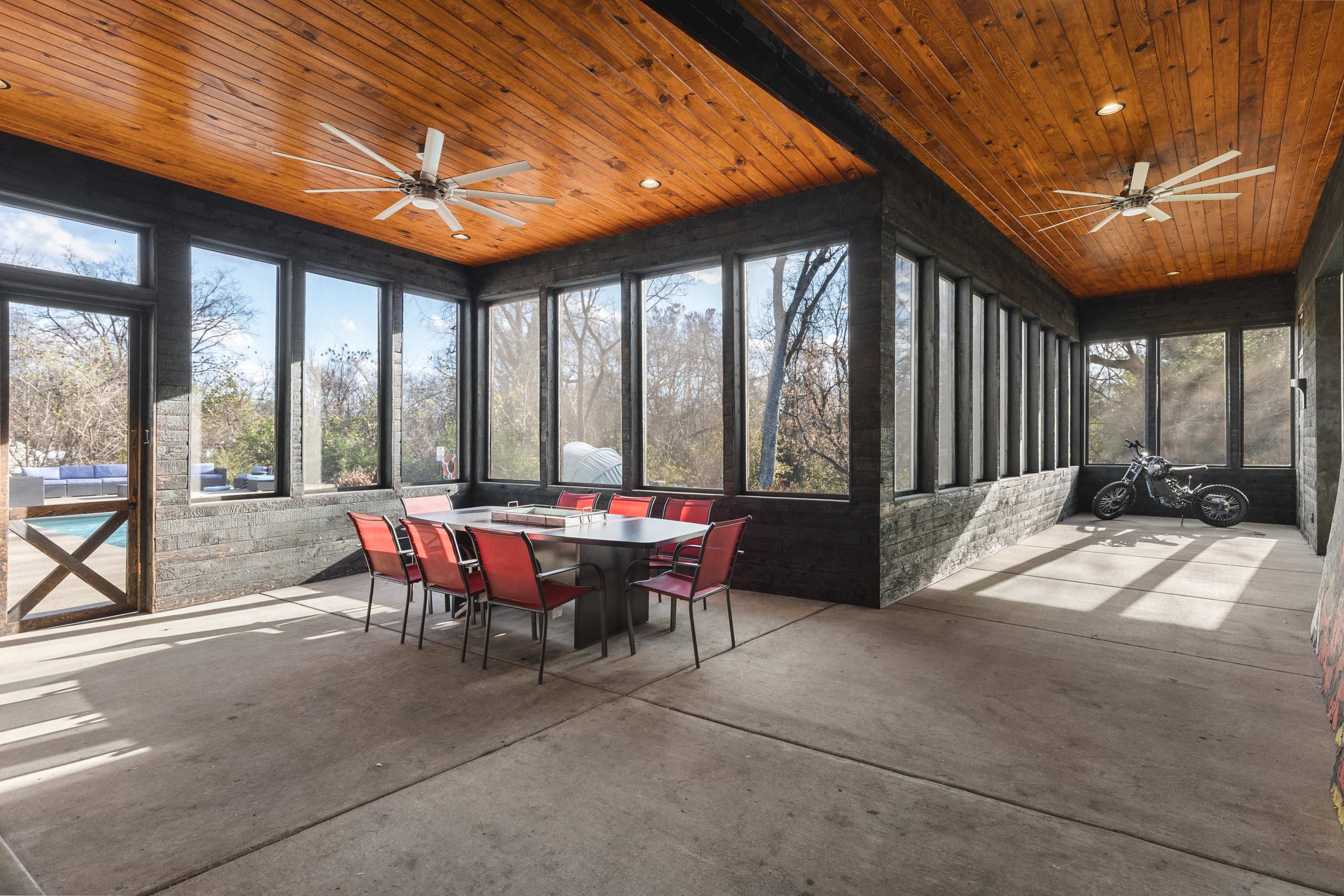 A screened-in porch featuring a large dining table with red chairs, wooden ceiling beams, and a motorcycle parked in one corner.