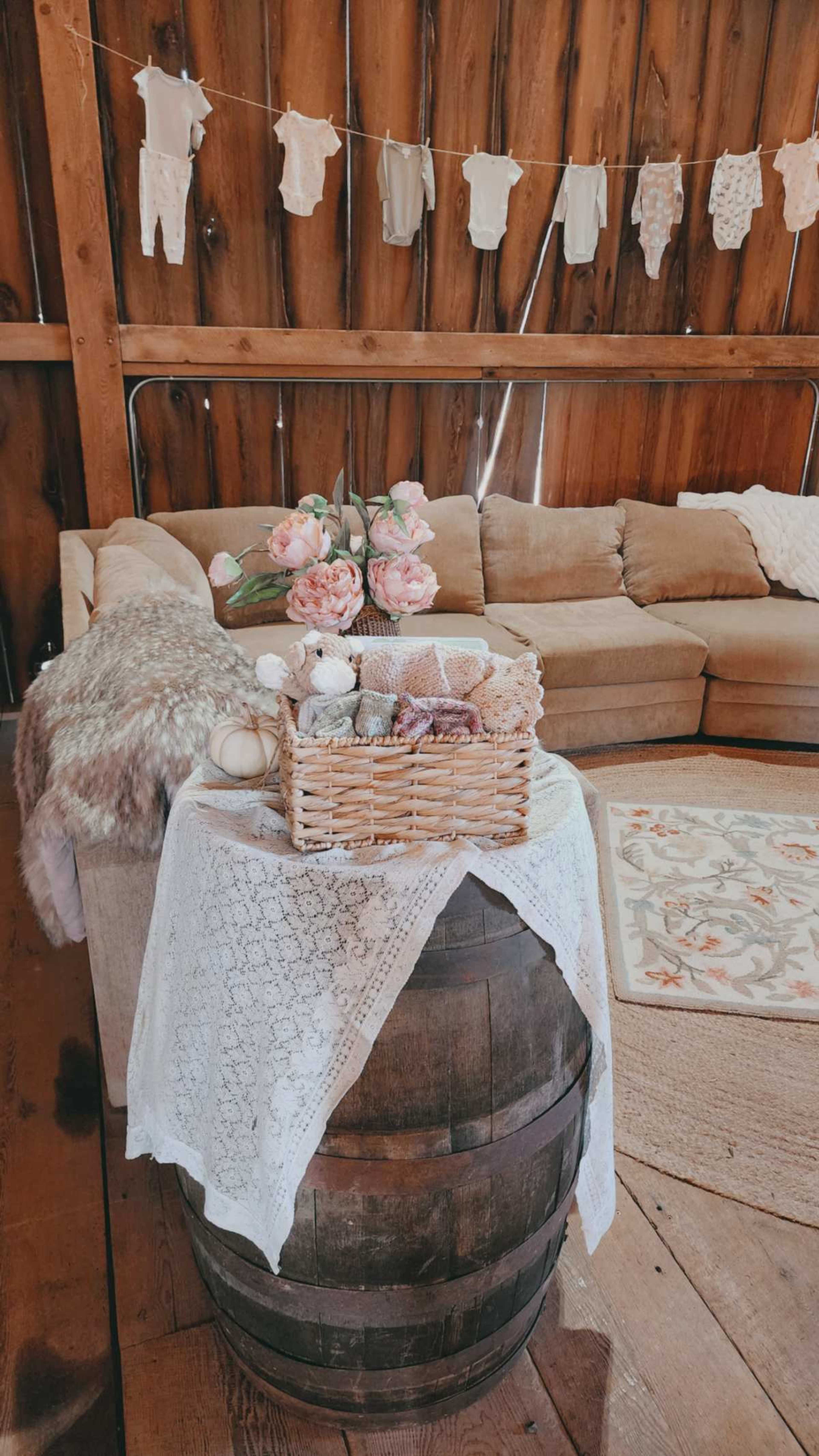 The scene depicts a cozy living area with a wooden barrel as a side table, adorned with lace and a basket of decorative items, against a backdrop of a rustic wooden wall and a beige sofa.