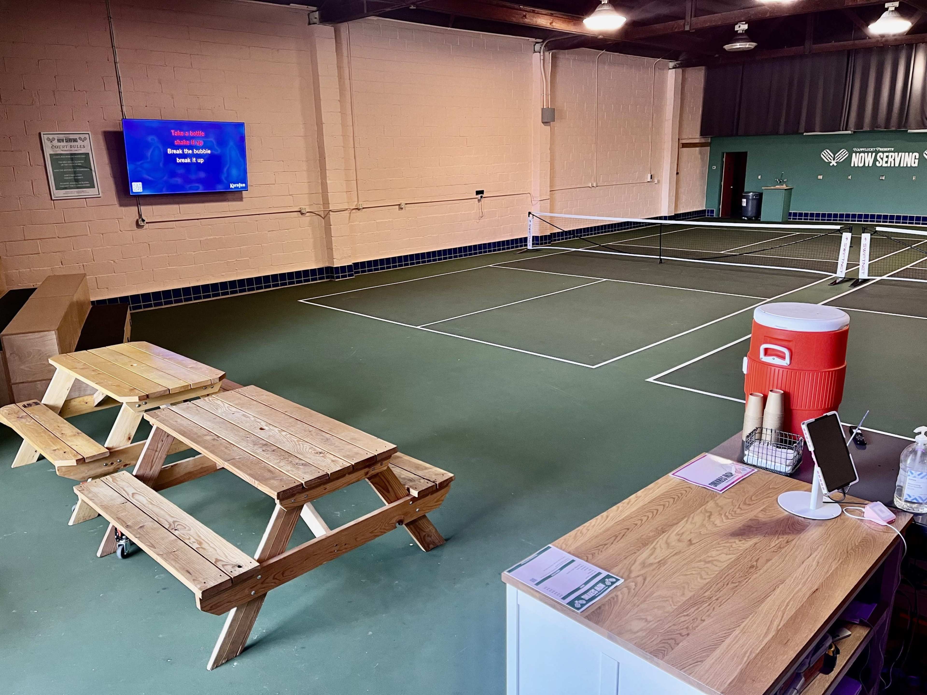 The image shows an indoor tennis facility with two courts, a picnic-style wooden table, a refreshment station, and a television displaying information on the wall.