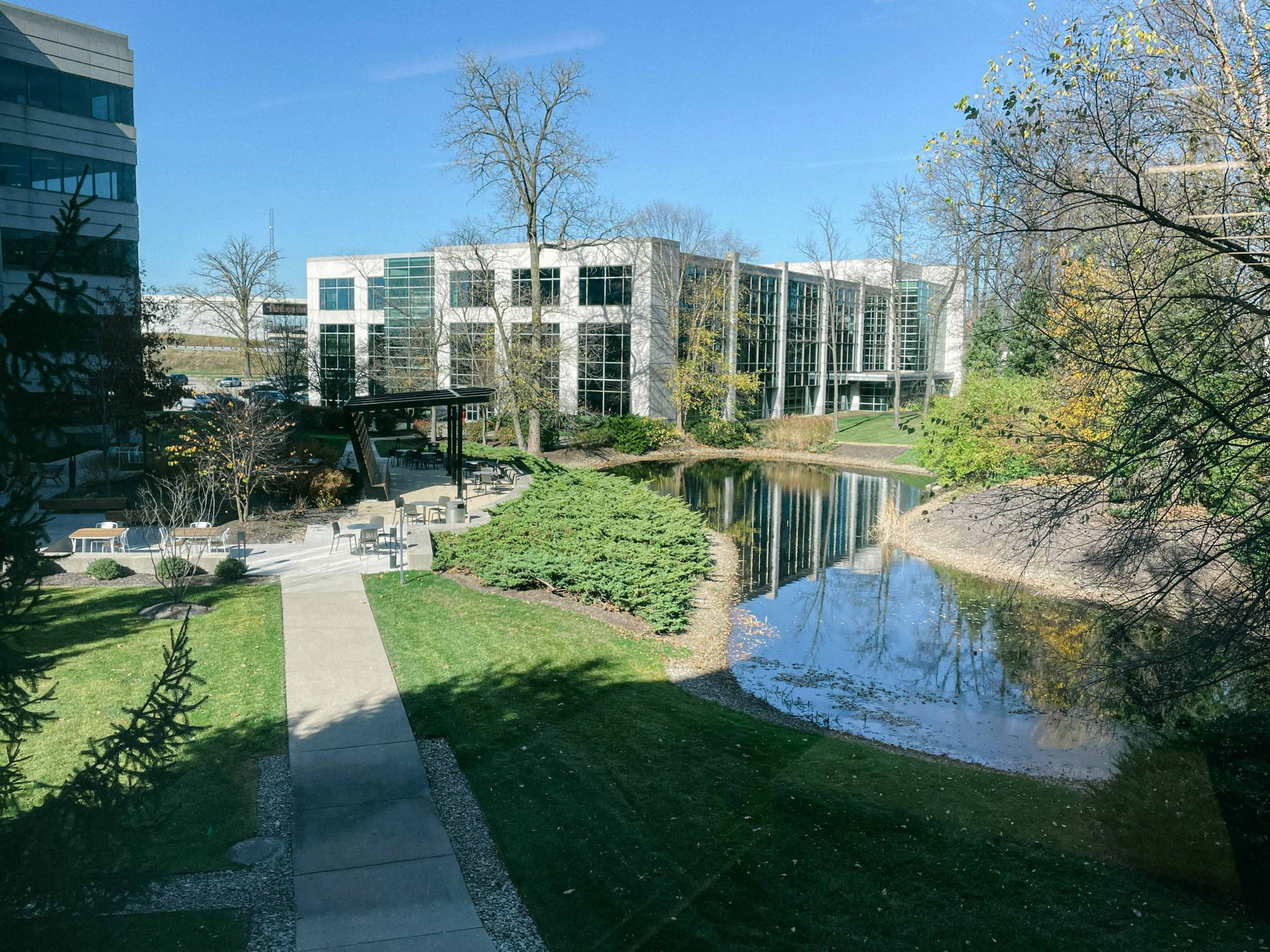 A landscaped area with a pond, surrounded by grass and trees, along with modern buildings in the background.