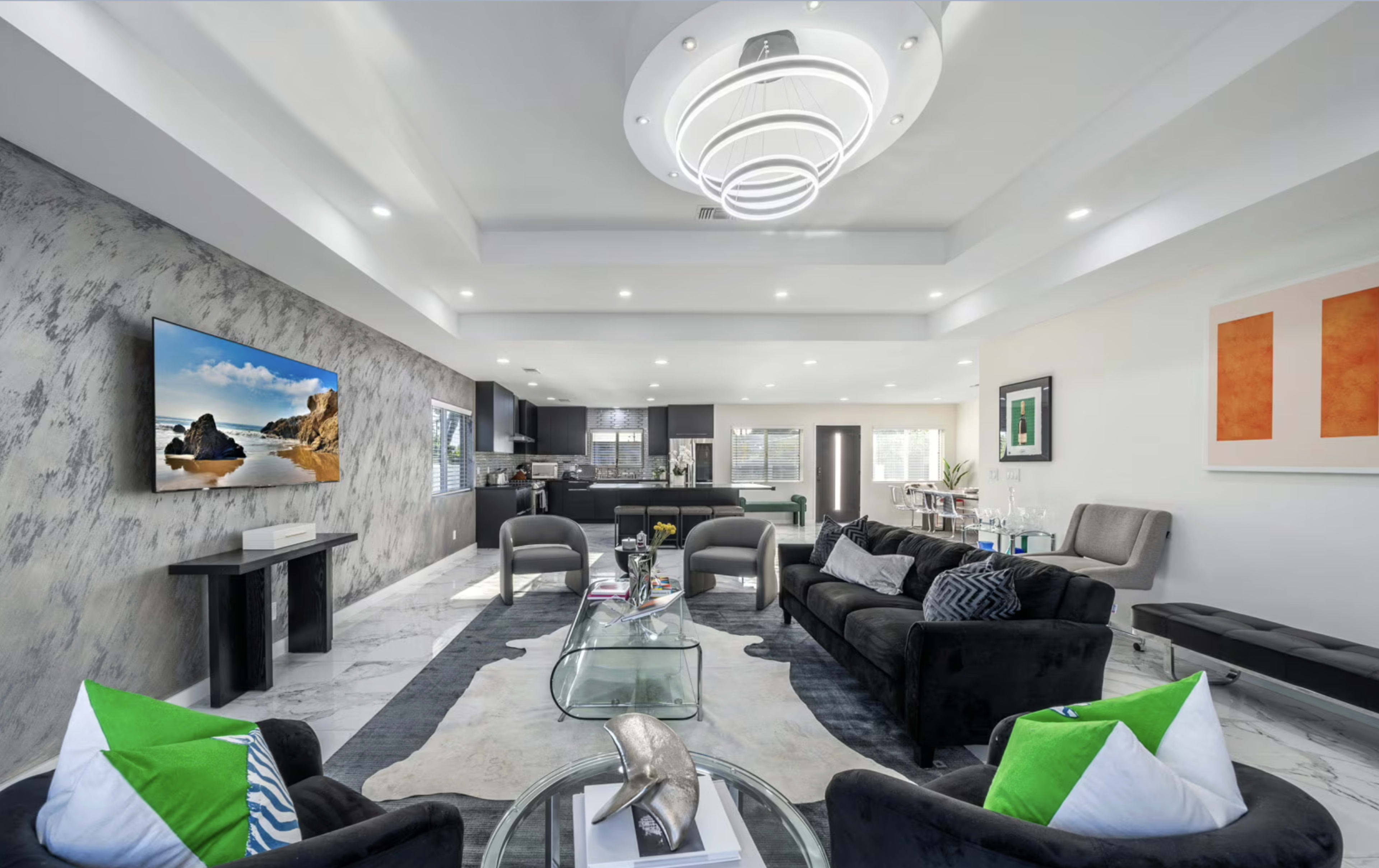 The image shows a modern living room featuring two black sofas, a glass coffee table, a large flat-screen TV mounted on the wall, and decorative pillows with green accents.