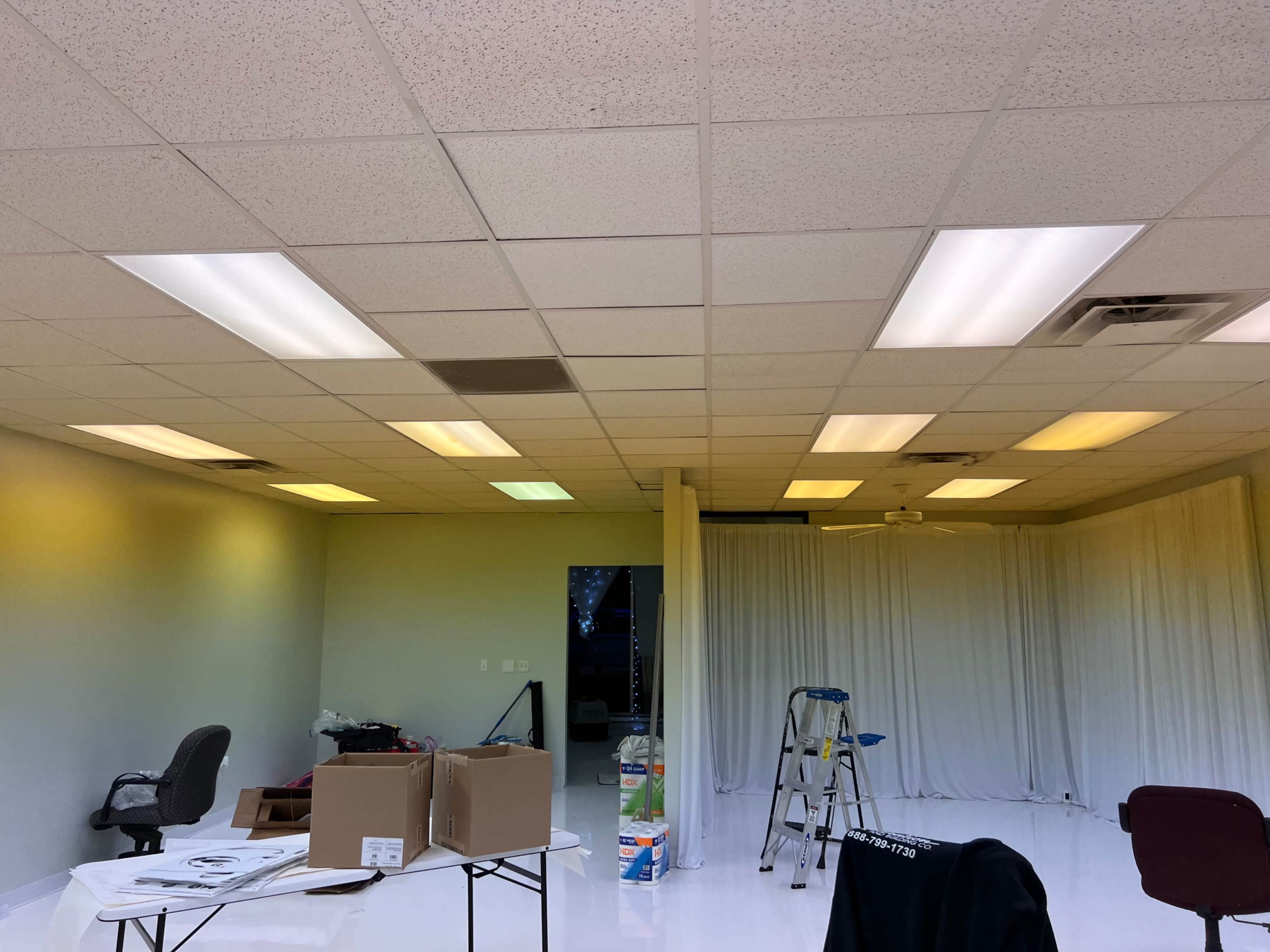 An empty room with fluorescent lighting, a ladder, some cardboard boxes, and a partially draped white curtain.