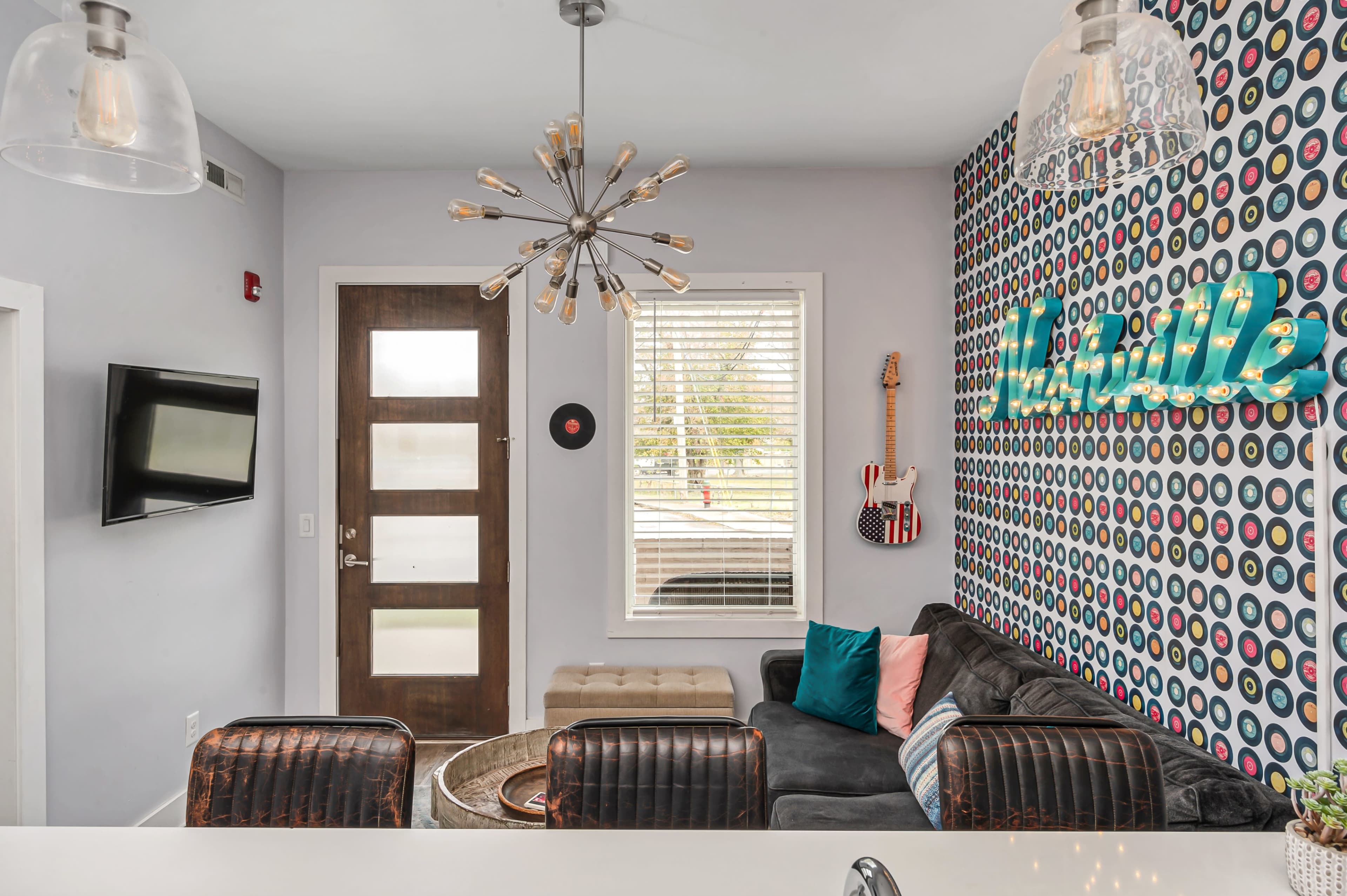 A living area with a gray couch, a decorative wall featuring colorful circular patterns, a television mounted on the wall, and a modern light fixture hanging from the ceiling.