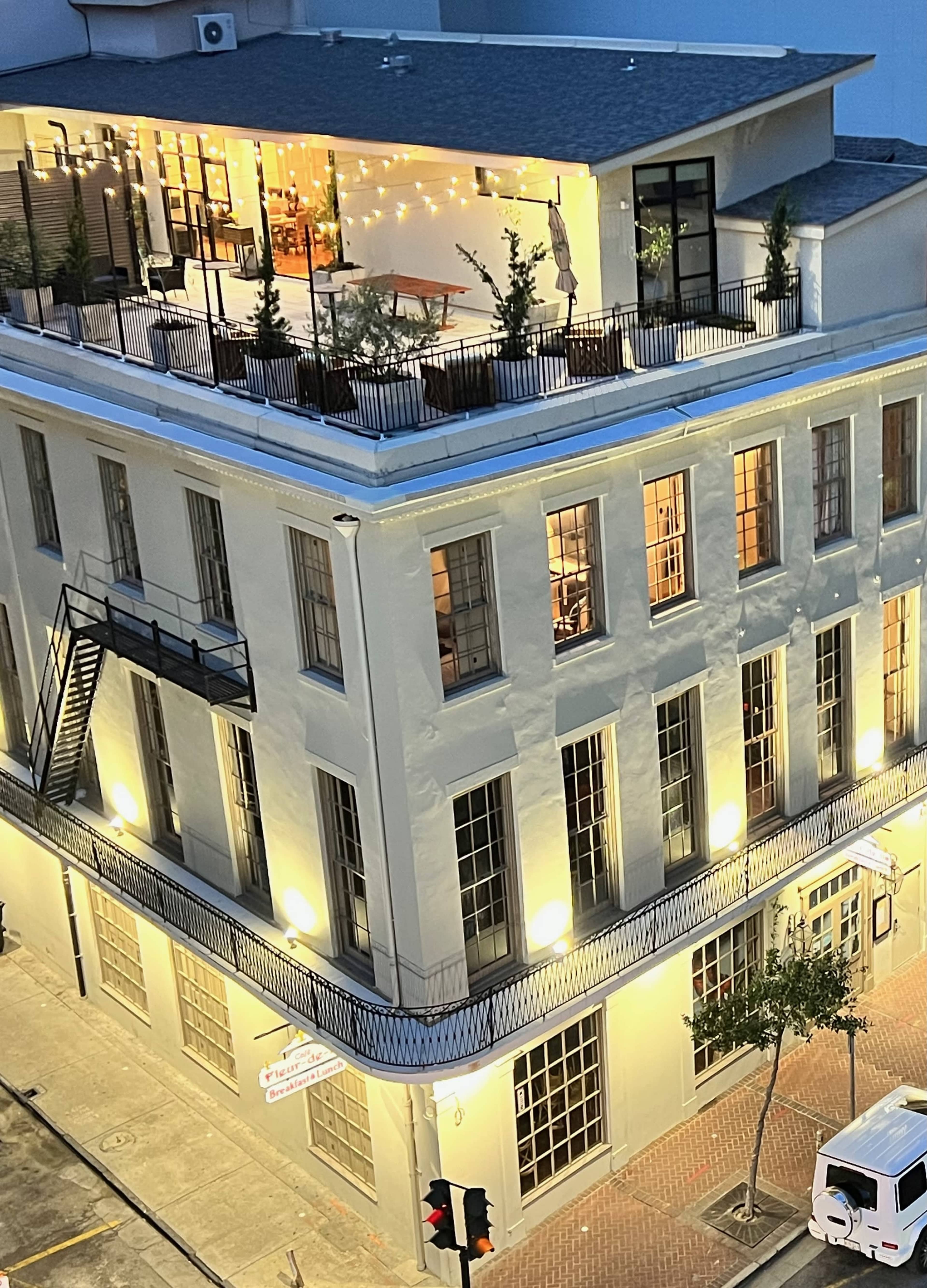 The image shows a multi-story building with a rooftop terrace adorned with string lights and potted plants, located at a street corner.