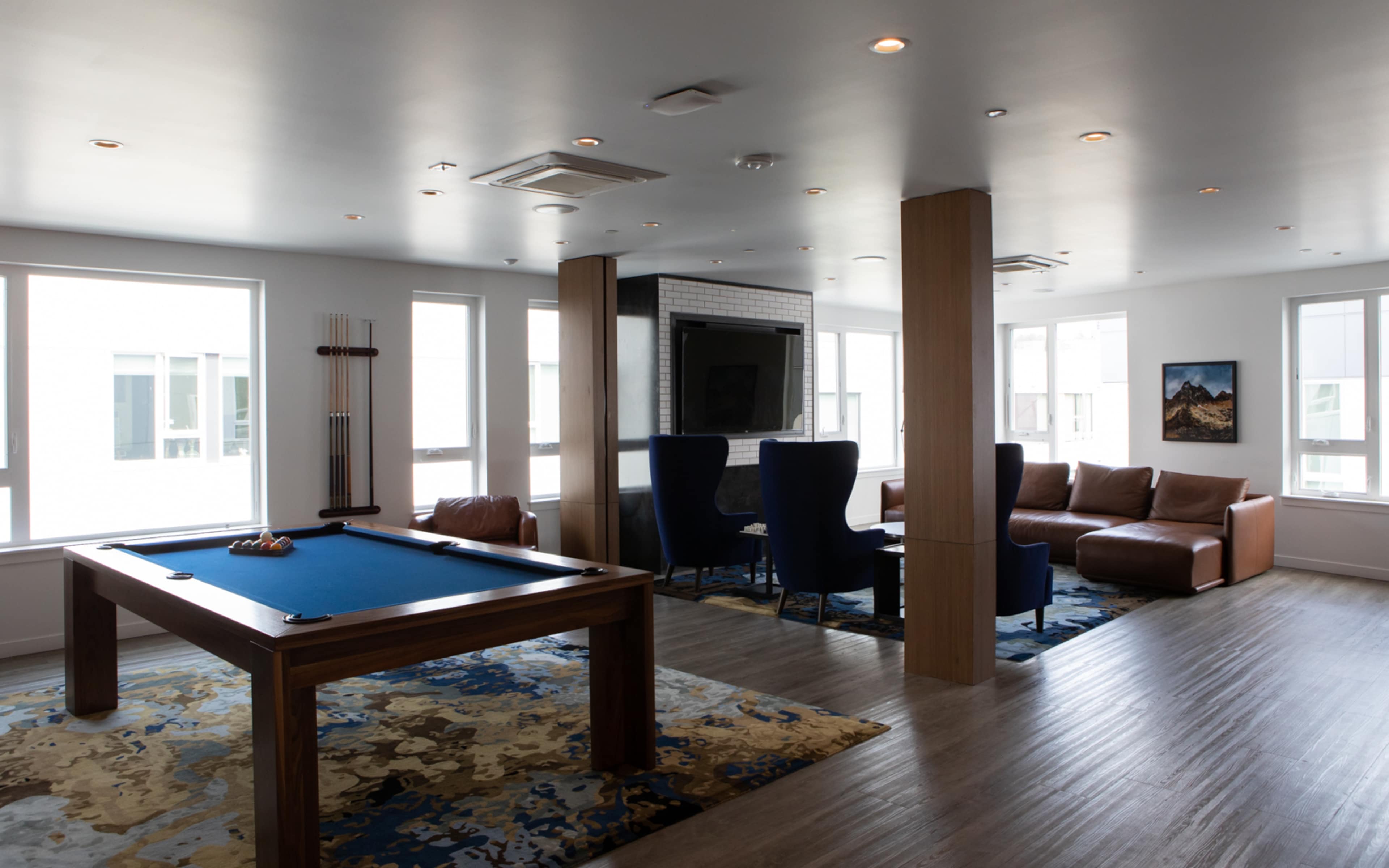 A modern game room featuring a pool table, a seating area with a leather couch, and large windows allowing natural light.
