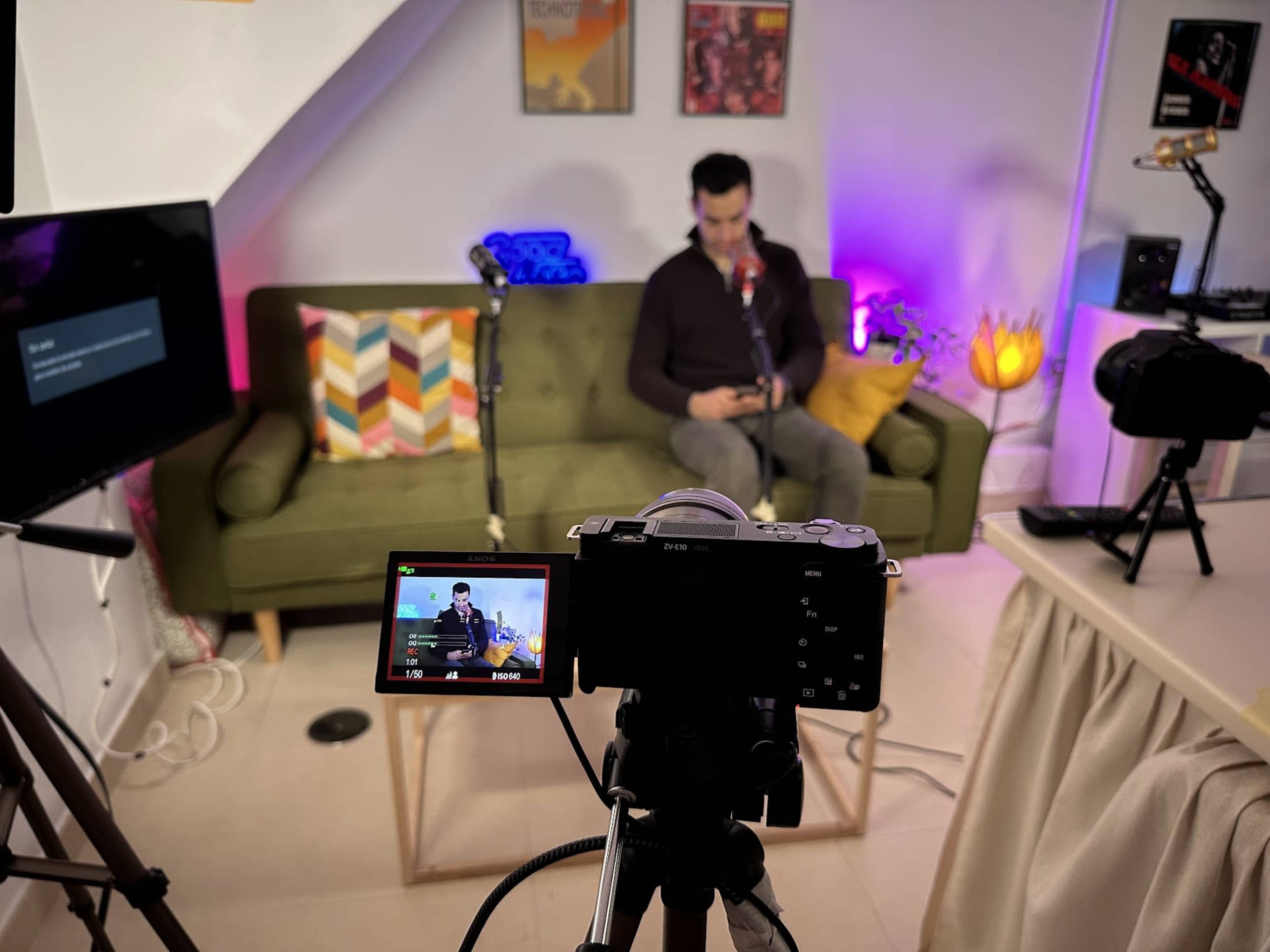 A camera is set up in a brightly lit room, focusing on a person sitting on a green couch while checking their phone, surrounded by microphones and colorful decorations.