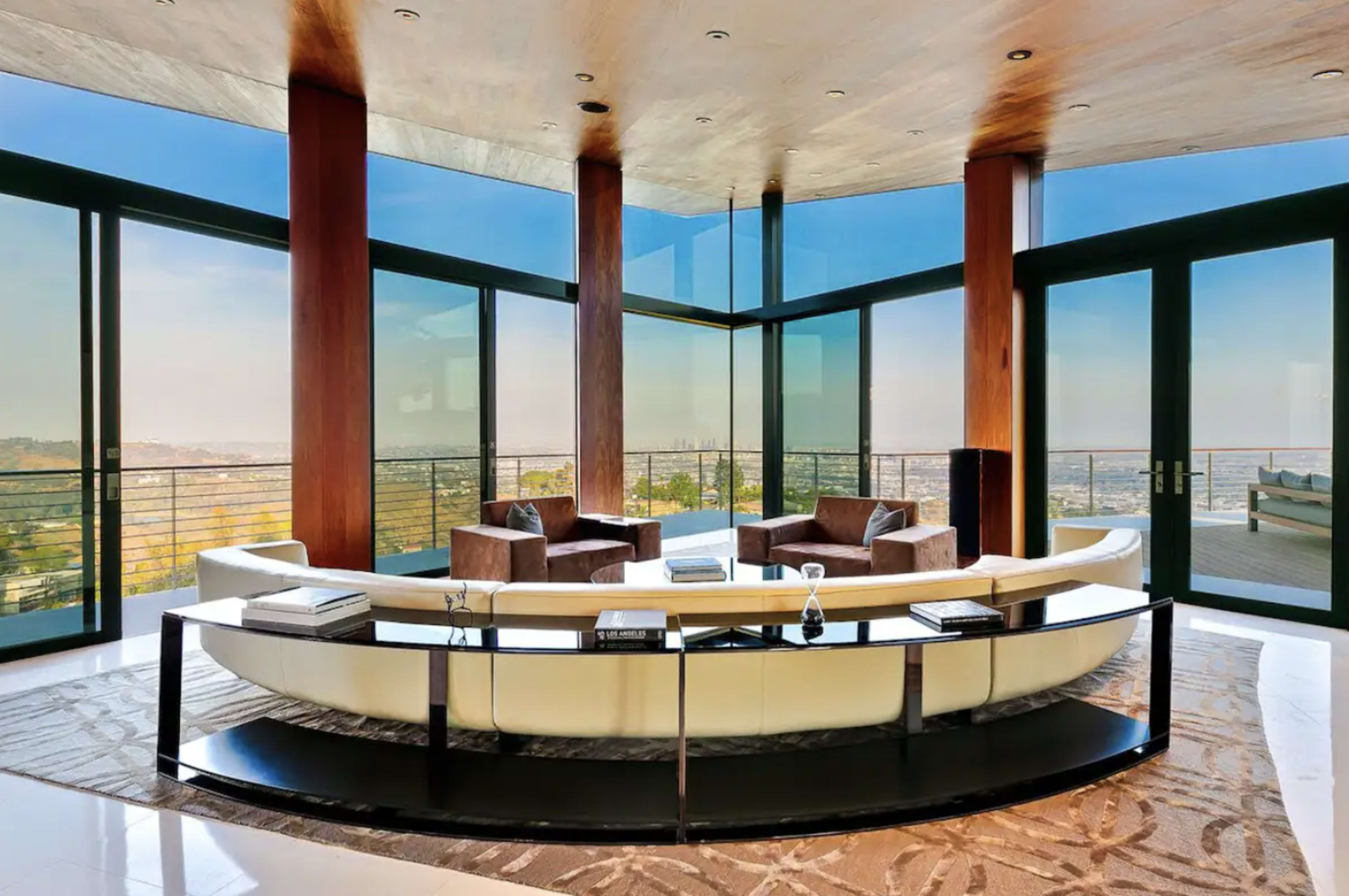 A modern living room features large glass windows with a panoramic view, a curved white sofa, and two brown armchairs.