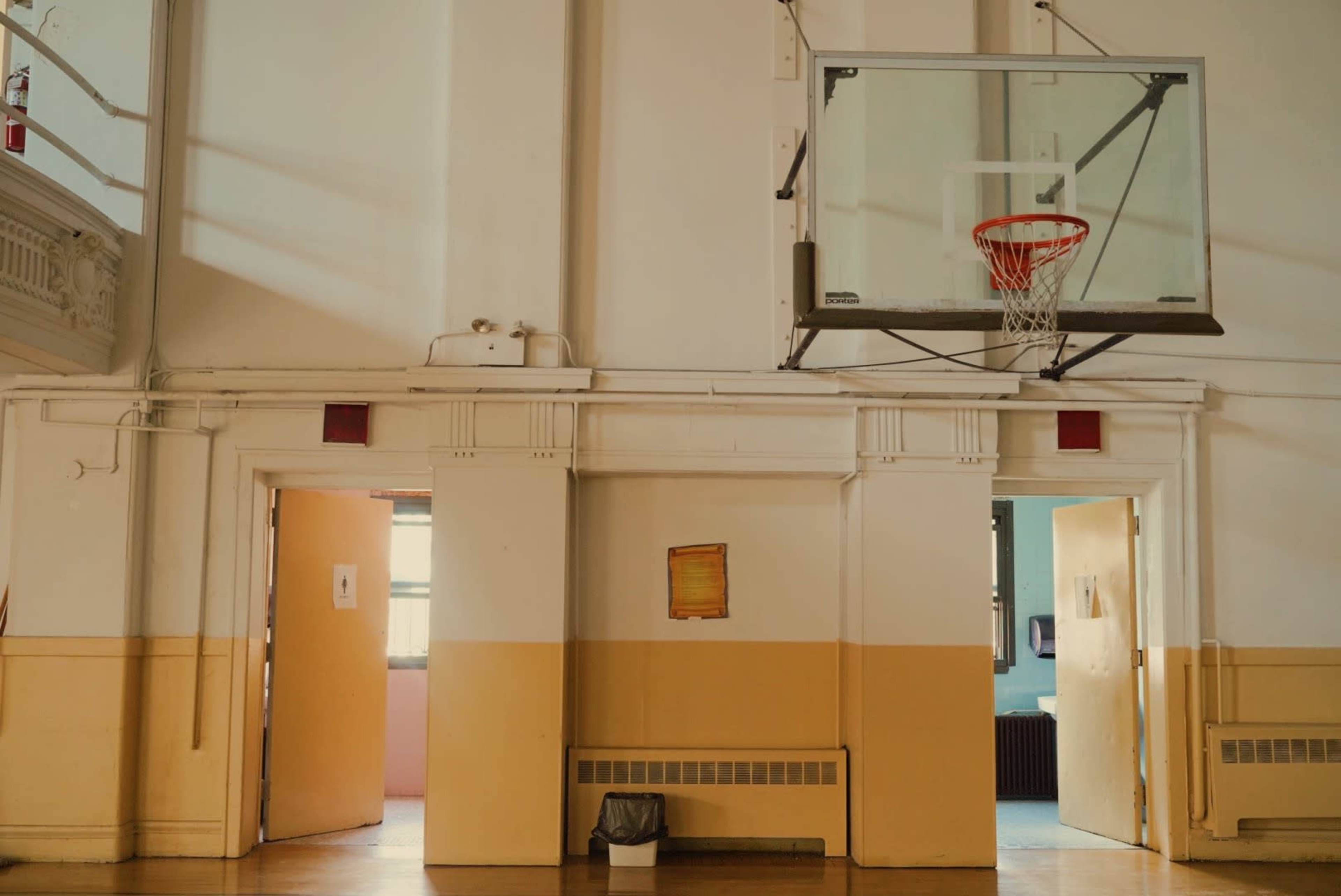 A gymnasium with a basketball hoop mounted on the wall, flanked by two open doorways.