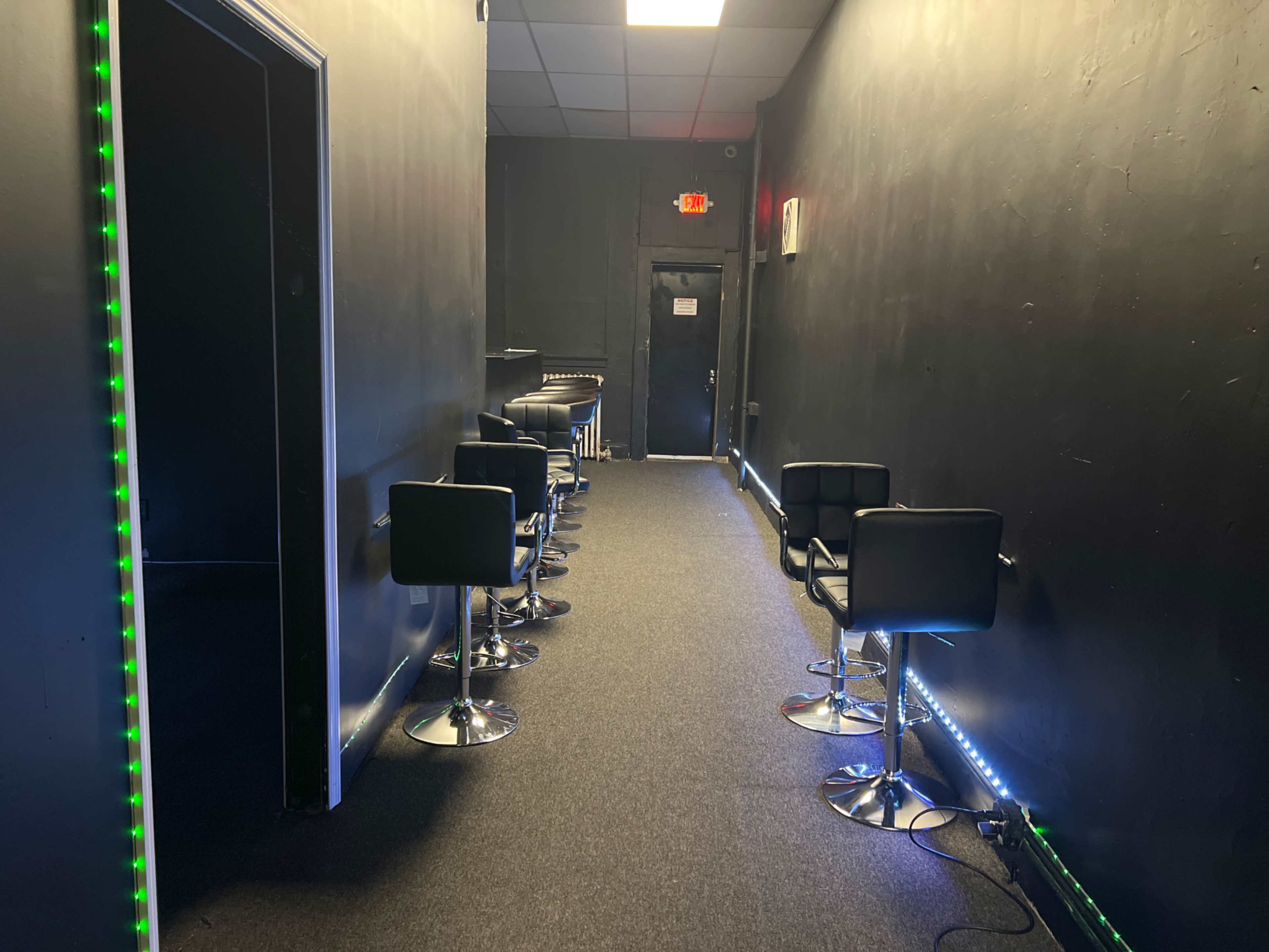 A narrow hallway features several black bar stools and dim lighting along the walls.