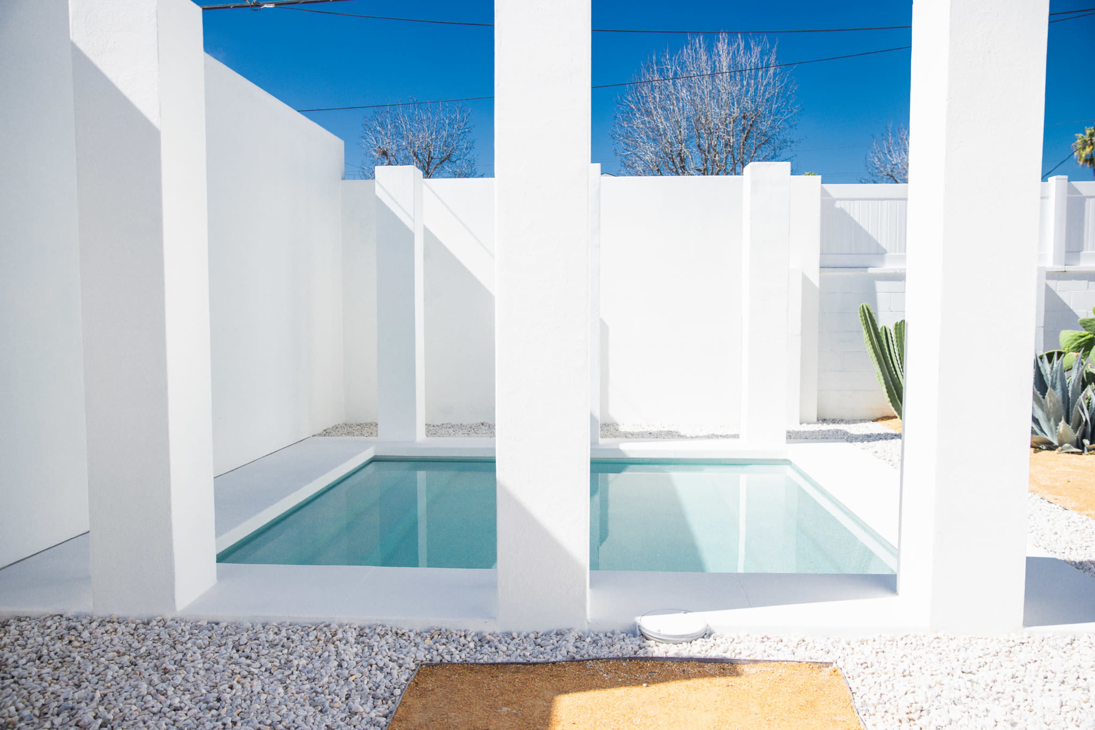 A minimalist outdoor space with a rectangular swimming pool surrounded by white walls, pebbled ground, and cacti.