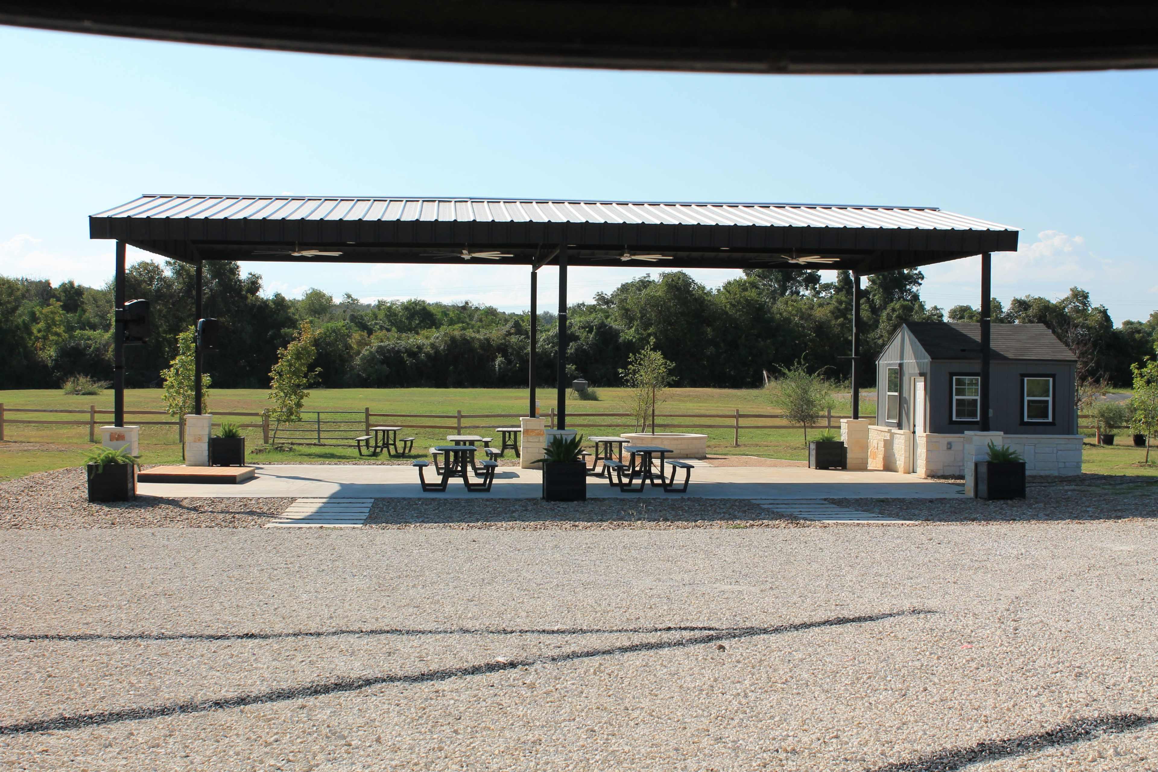 The image shows a covered outdoor pavilion with picnic tables and a small kiosk, set against a backdrop of greenery.