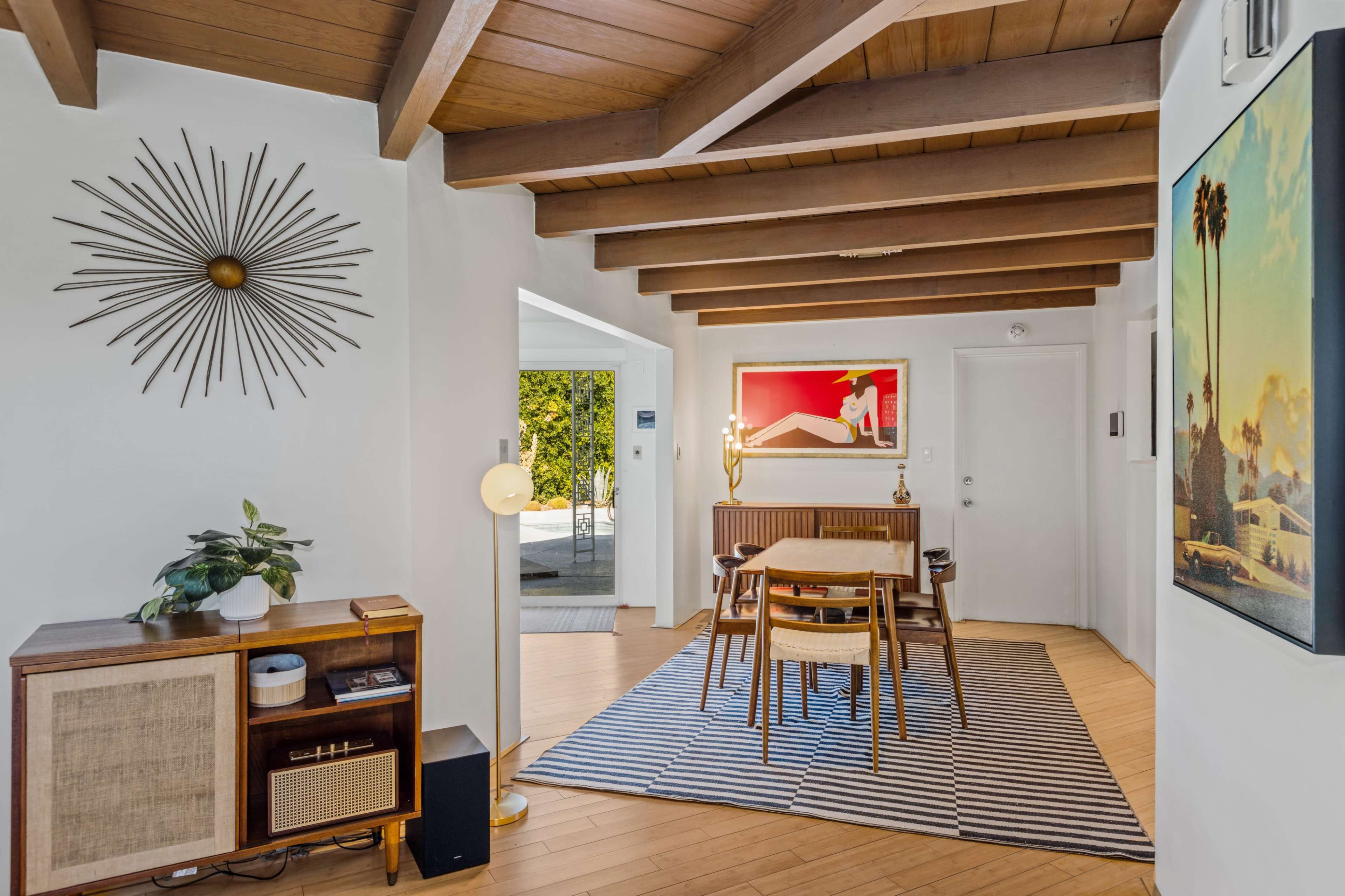 A modern dining area features a wooden table surrounded by chairs, with a mid-century style sideboard, a starburst wall decor, and a large artwork on the adjacent wall.