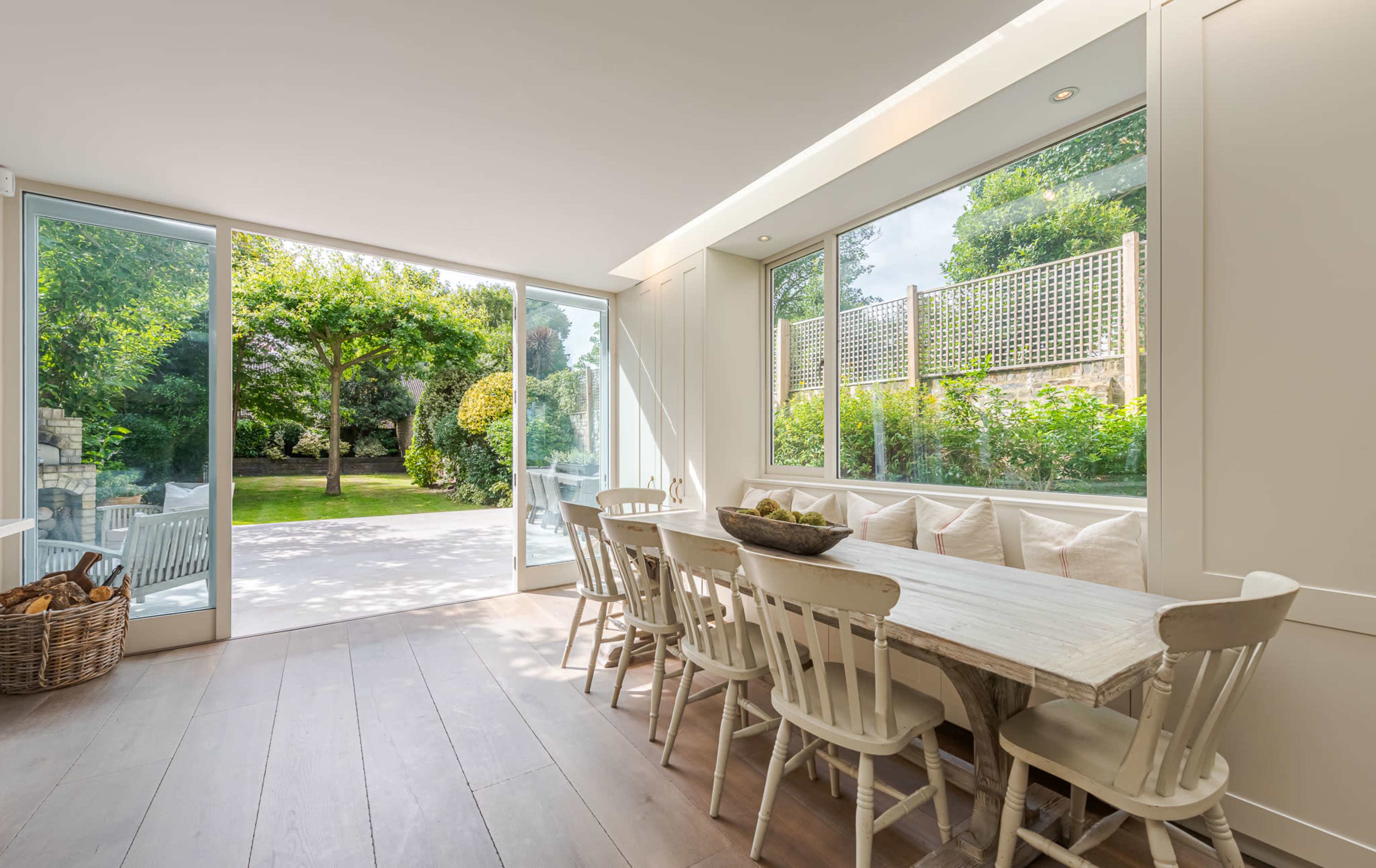 A spacious dining area features a long white table surrounded by wooden chairs, with large glass windows showcasing a garden view.