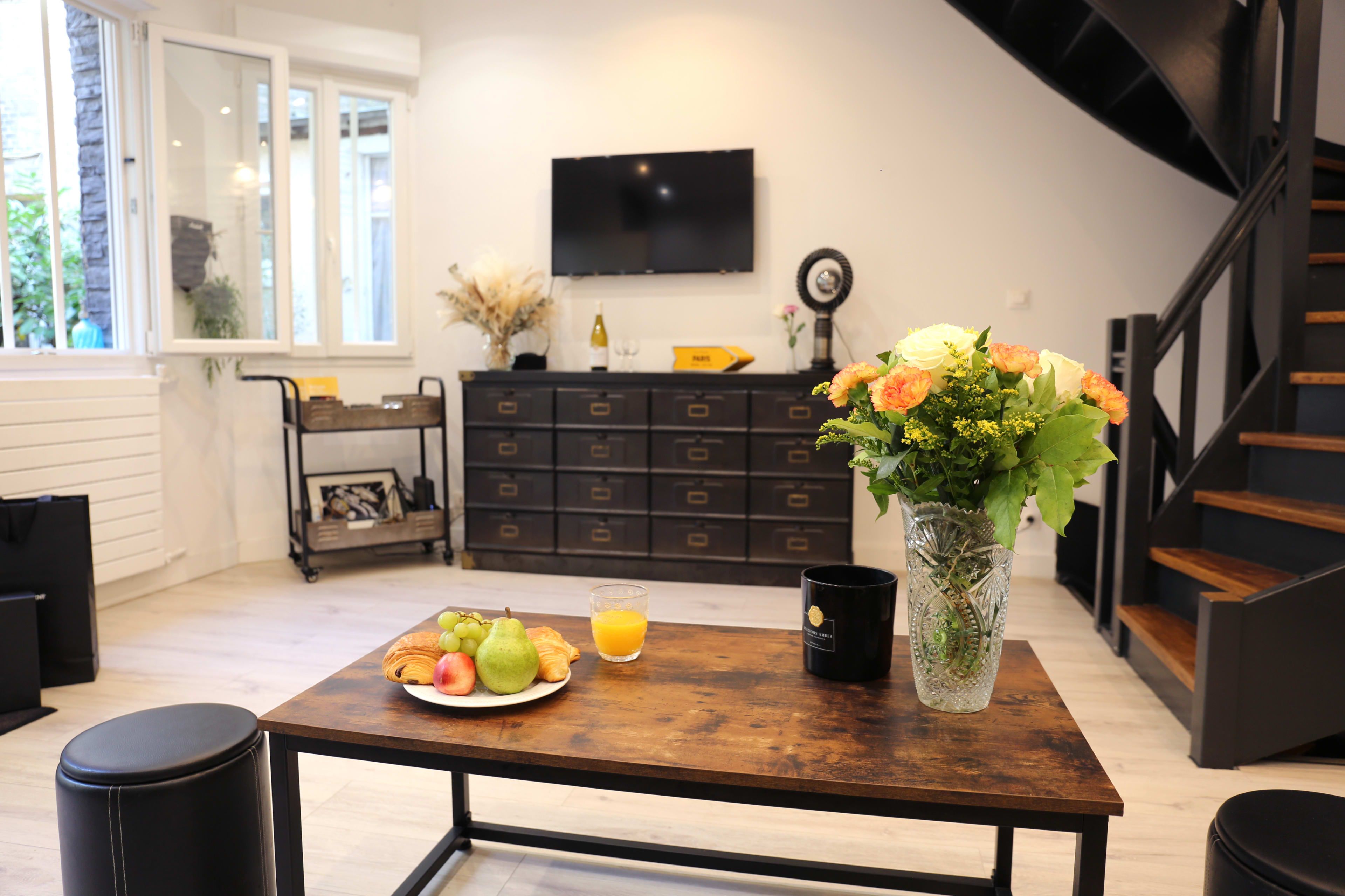 A wooden coffee table holds a plate of croissants, fruits, and a glass of orange juice in a modern living room with a staircase and a television.