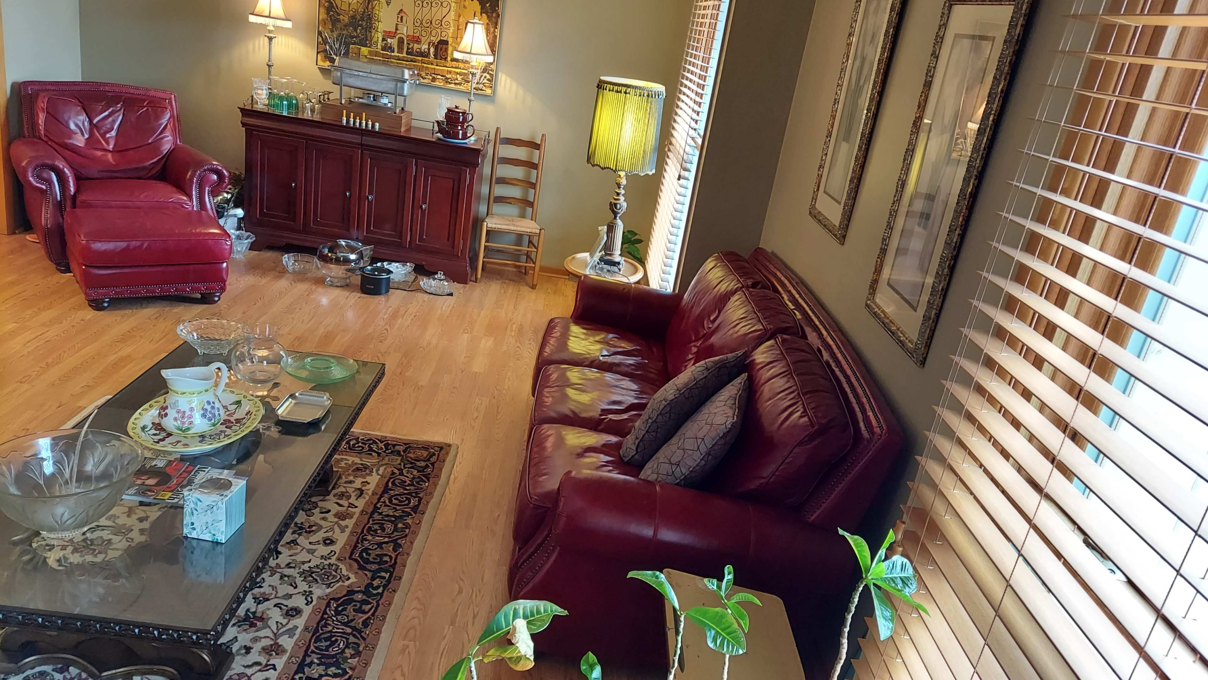A living room featuring a red leather sofa and armchair, a coffee table with various items, and decorative lamps alongside wooden furniture.