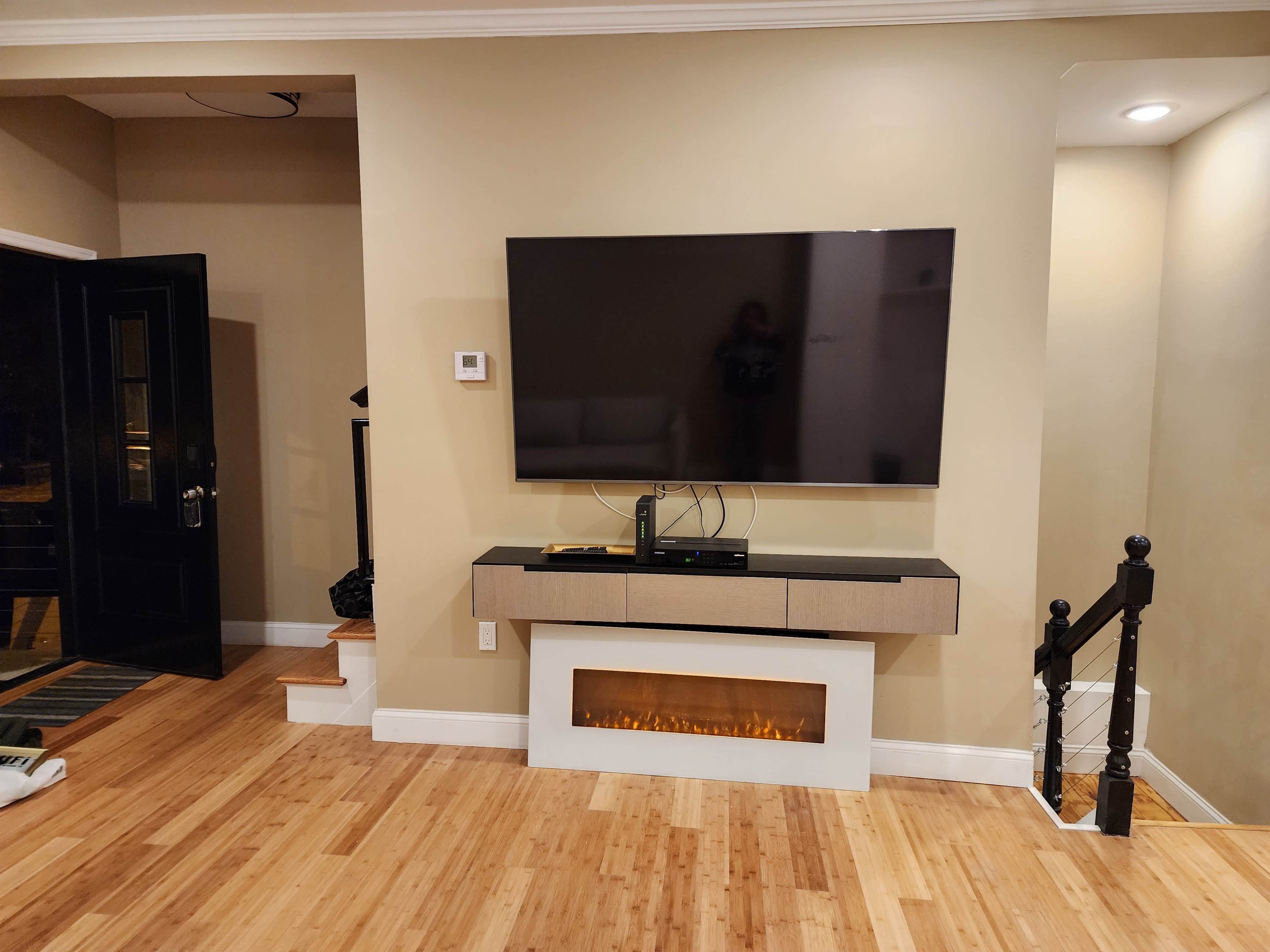 A wall-mounted television is positioned above a sleek console with an electric fireplace, adjacent to a staircase and entrance door in a room with light-colored walls and a bamboo floor.