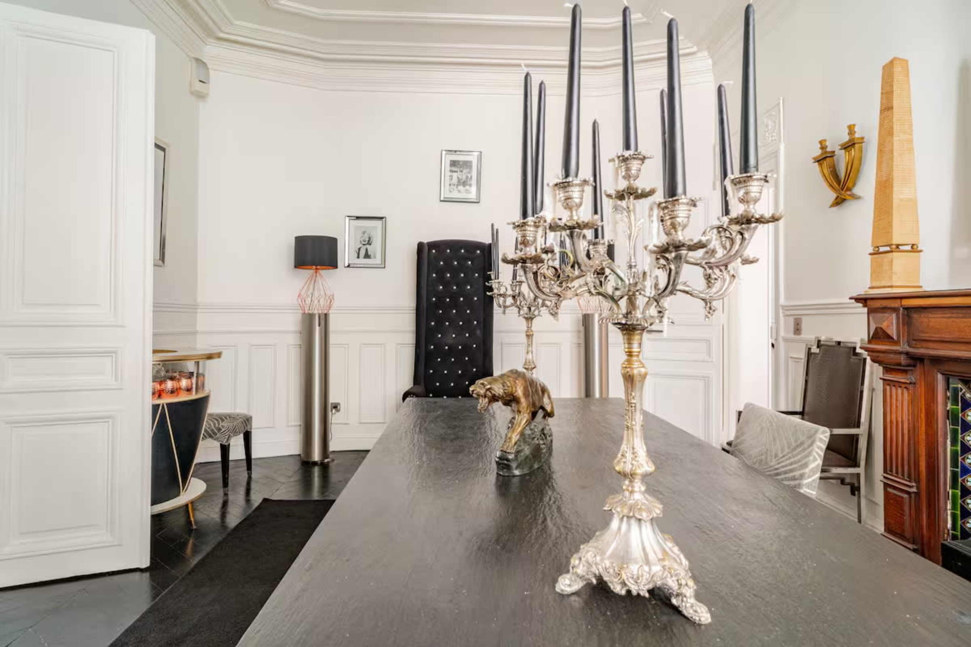 The image shows a dining room with a long black table, a silver candelabra with black candles, a tall black lamp, and various chairs arranged around the table.