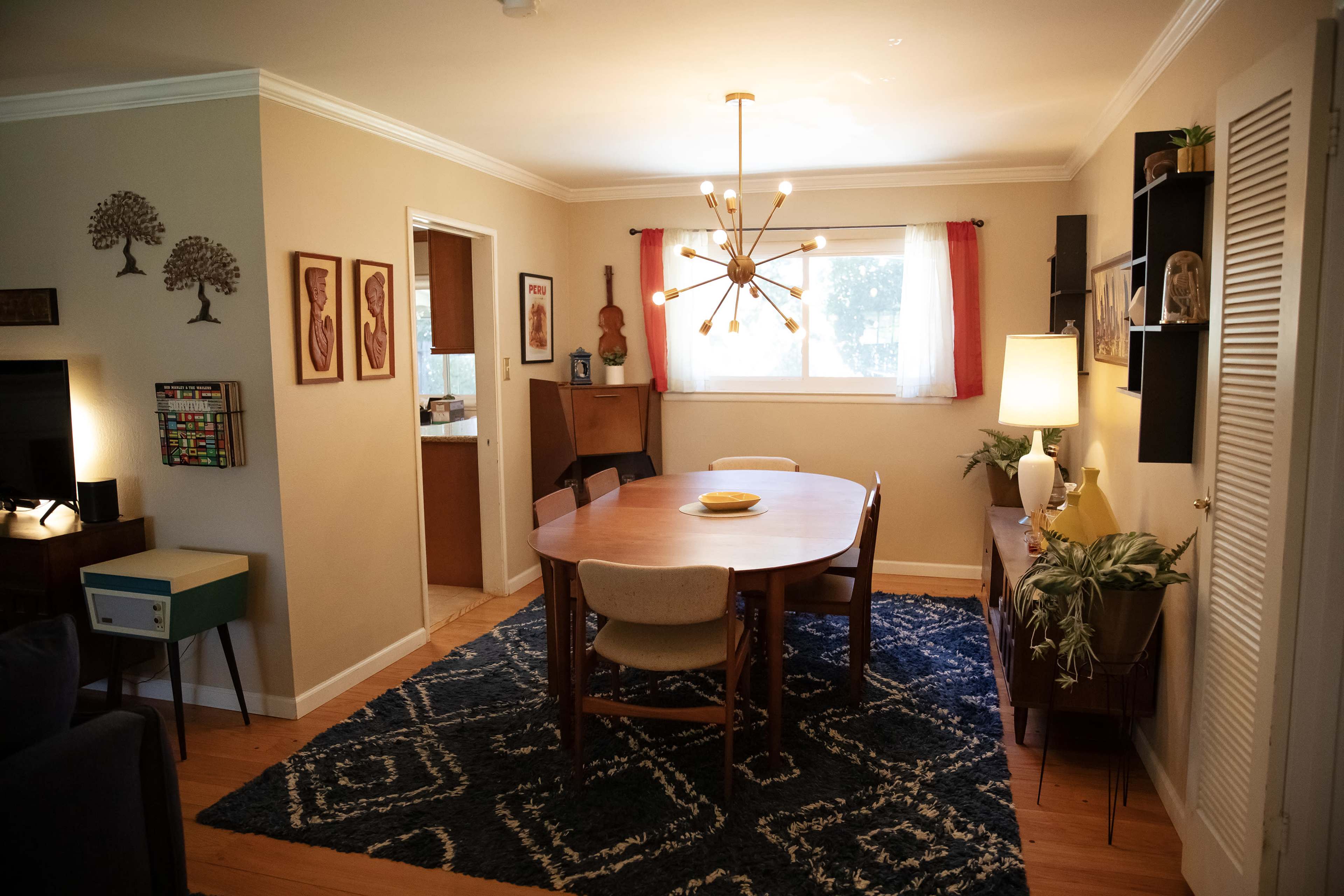 A dining area features a round wooden table surrounded by chairs, with a large window, pendant light, and decorative elements on the walls.