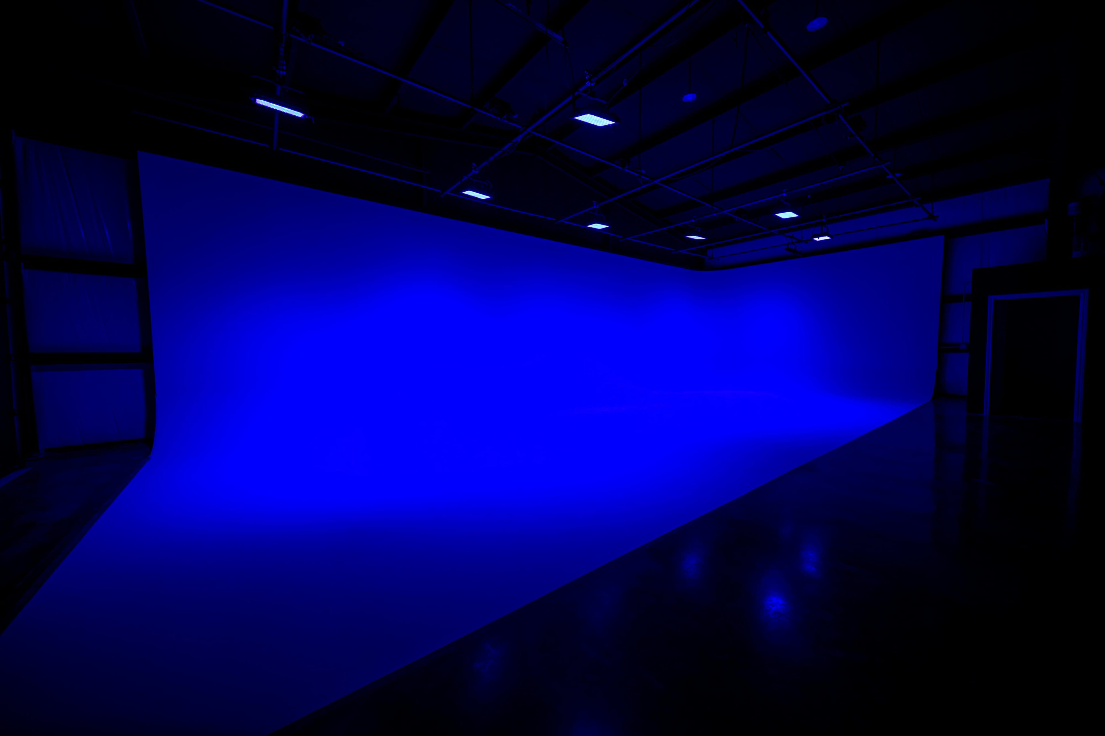 The image shows a spacious indoor studio with a large blue backdrop and polished dark flooring, illuminated by overhead lights.