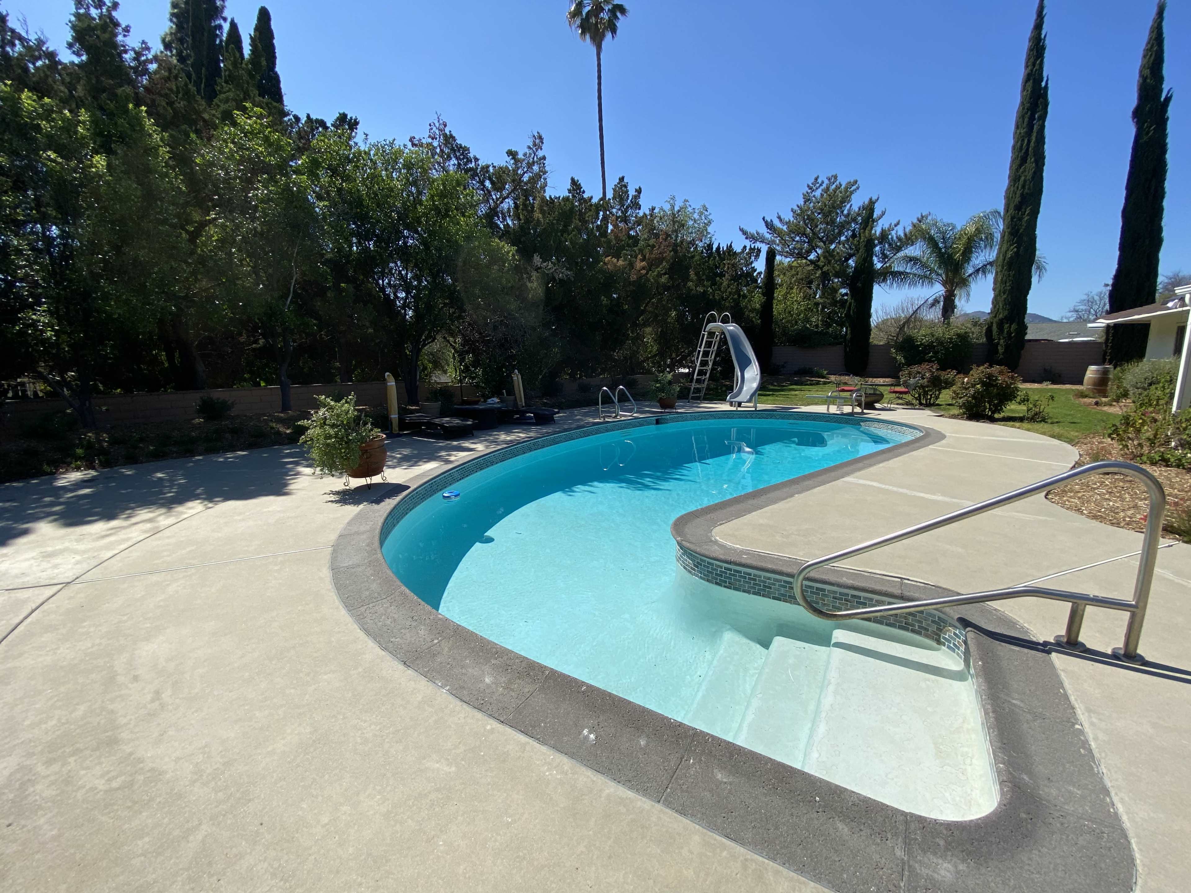 A curved swimming pool with a slide, surrounded by trees and lounge chairs on a sunny day.