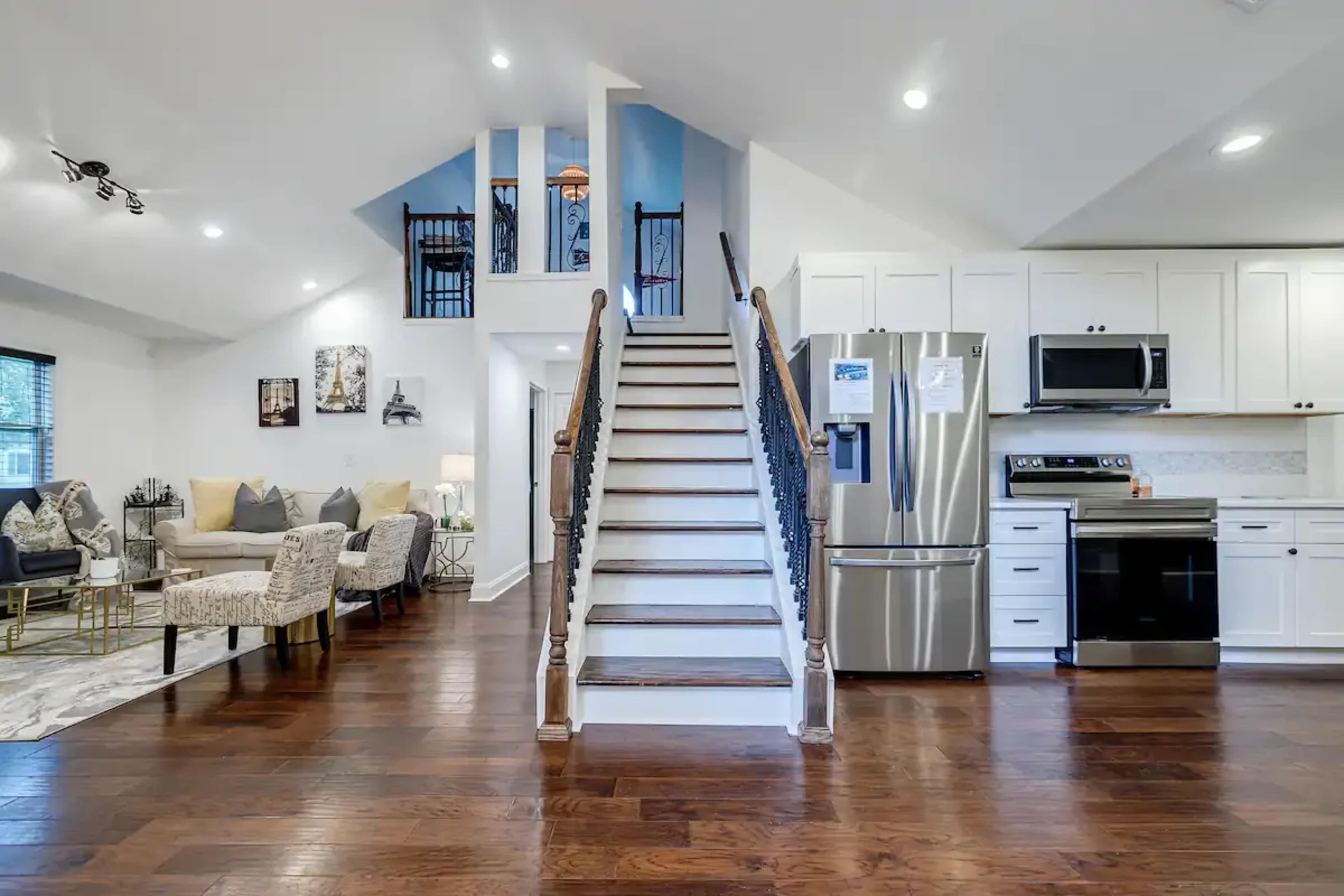 A spacious, modern living room connected to a kitchen area, featuring a staircase with wooden steps and a wrought iron railing.