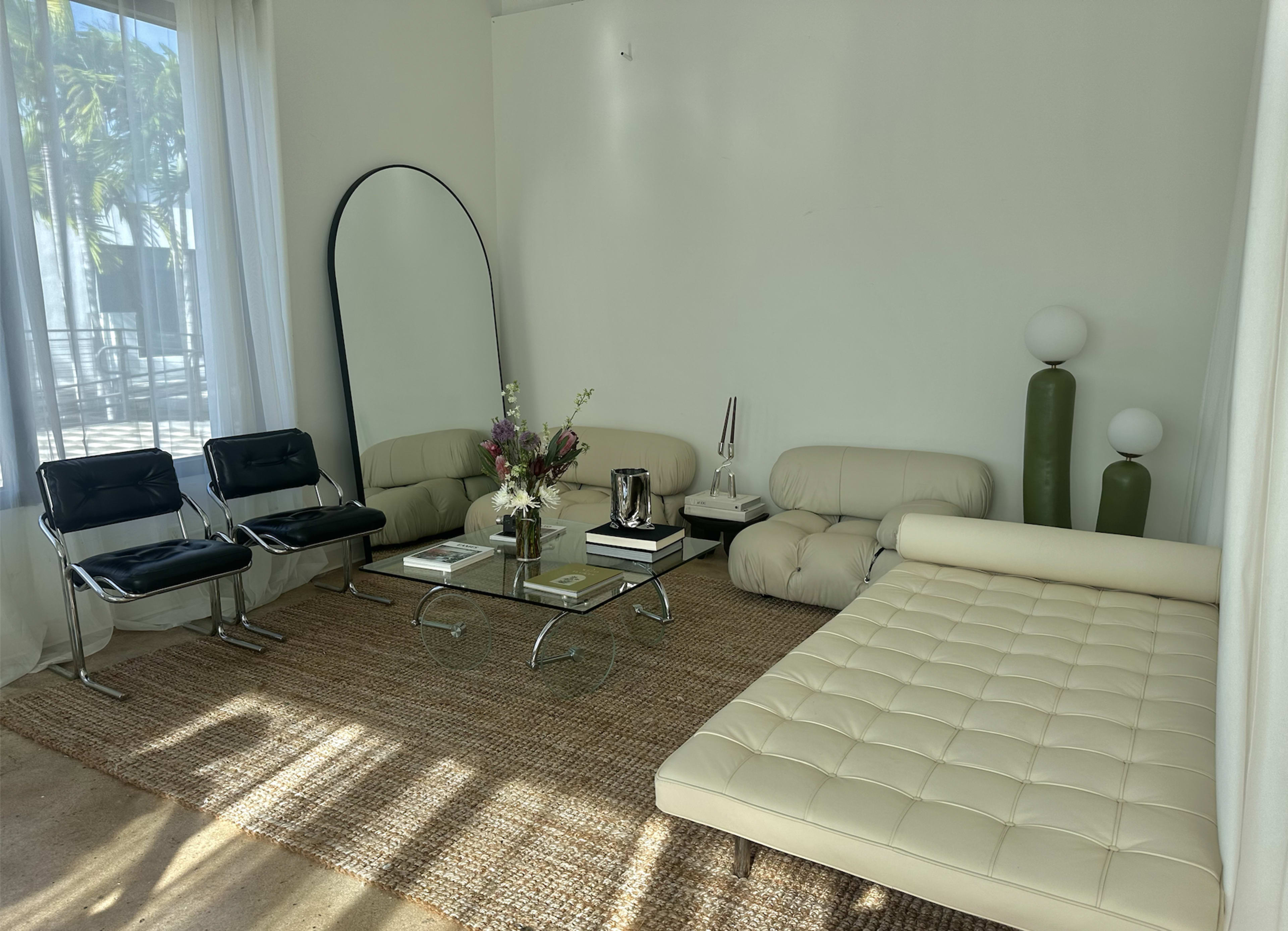 The image shows a modern living room with a light color scheme, featuring a glass coffee table, two leather sofas, and decorative vases.