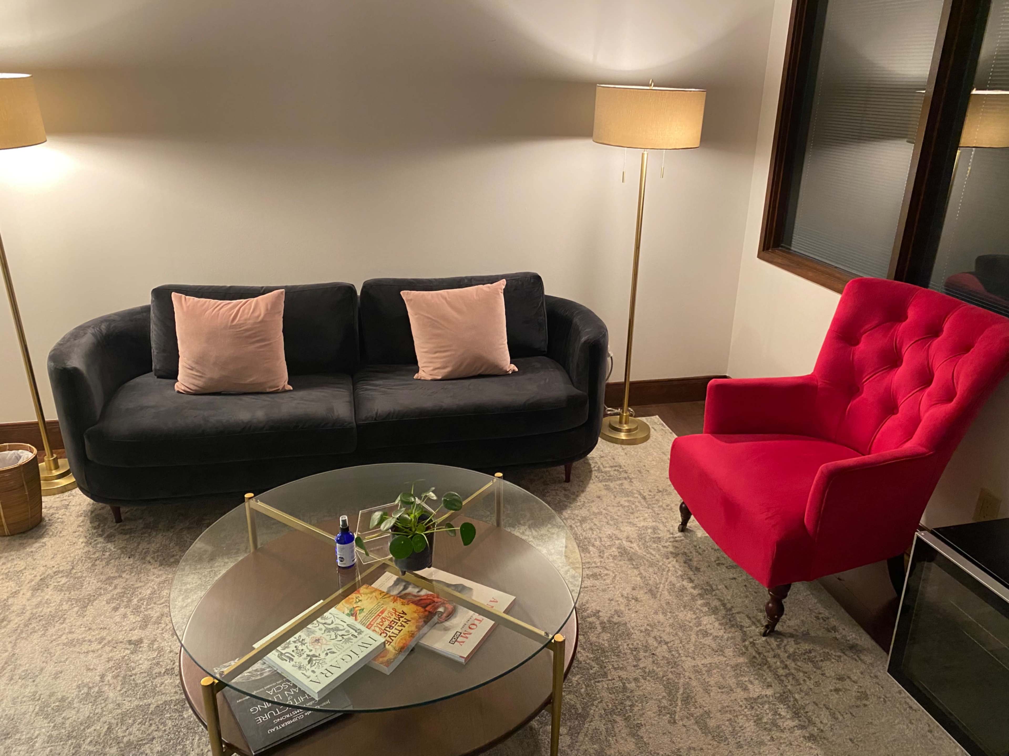 A black sofa with pink cushions faces a red armchair, alongside a round glass coffee table with various magazines and a small plant.