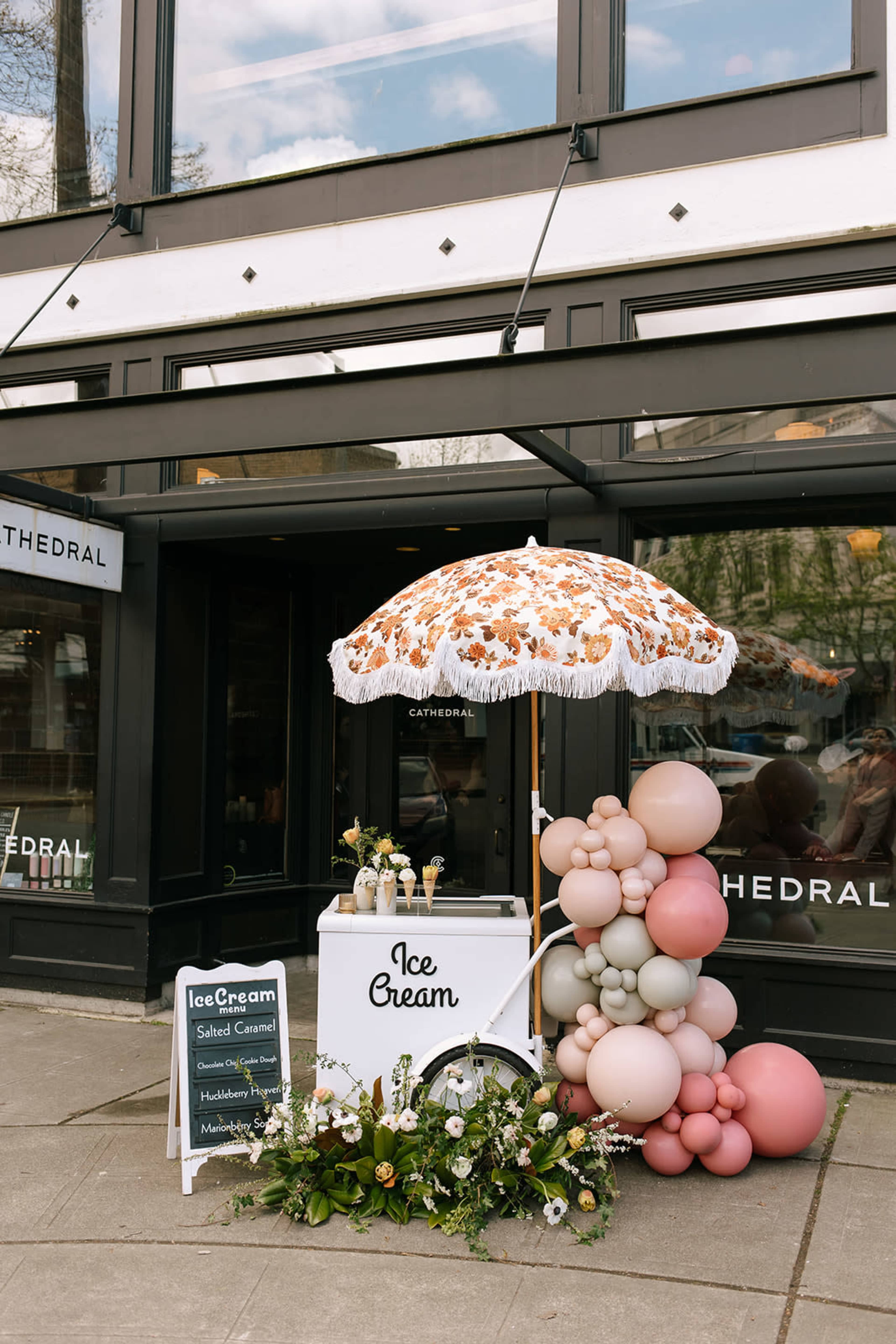 An ice cream cart with a decorative umbrella and a balloon display is set up outside a building with large windows.