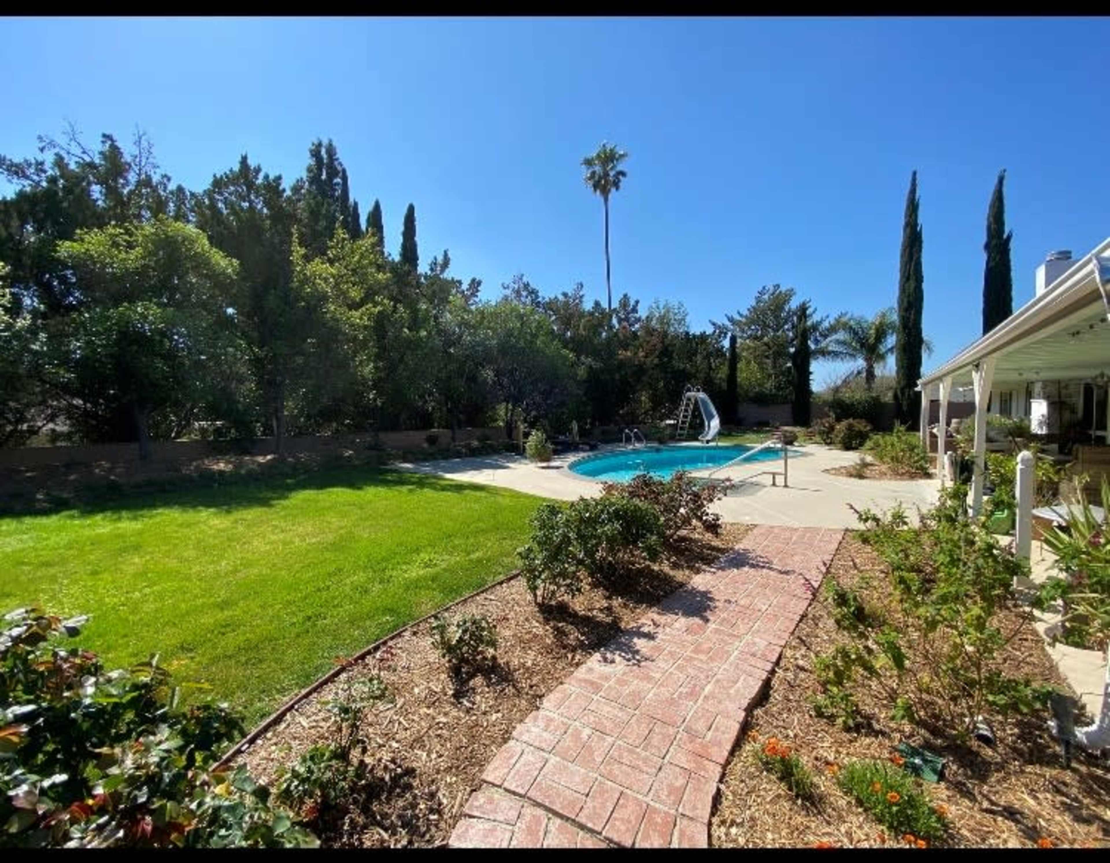 A backyard features a swimming pool surrounded by grass, palm trees, and landscaped flower beds.
