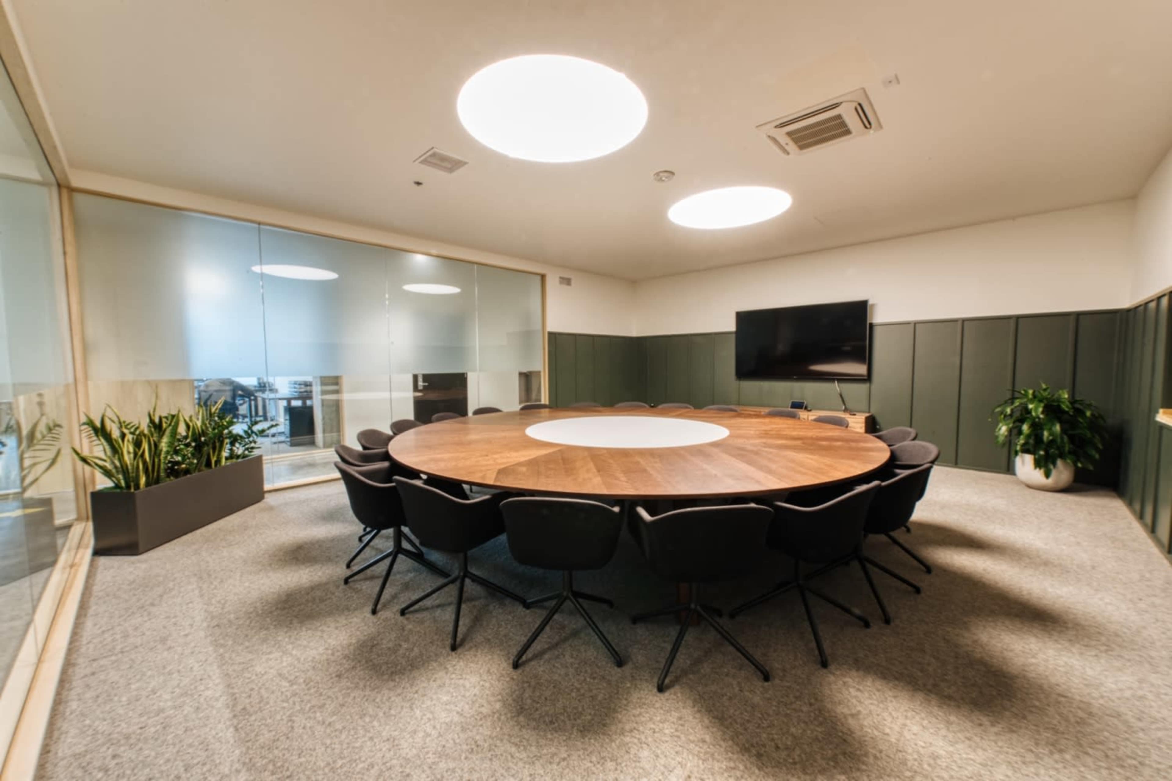 The image shows a modern conference room featuring a circular wooden table surrounded by black chairs, with glass walls and a large screen on one side.