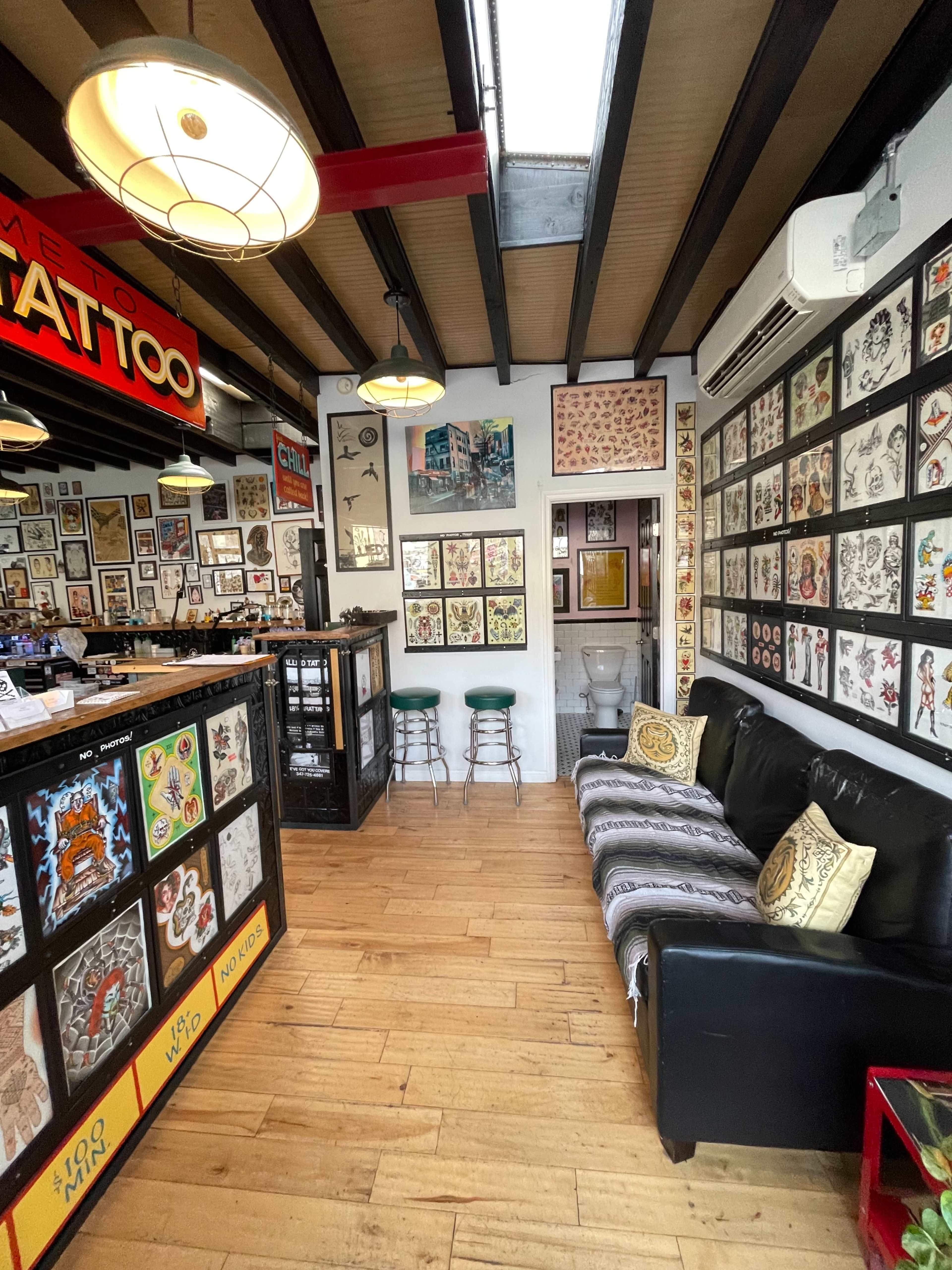 The interior of a tattoo parlor features wooden beams, a black leather couch, and walls adorned with various tattoo designs.