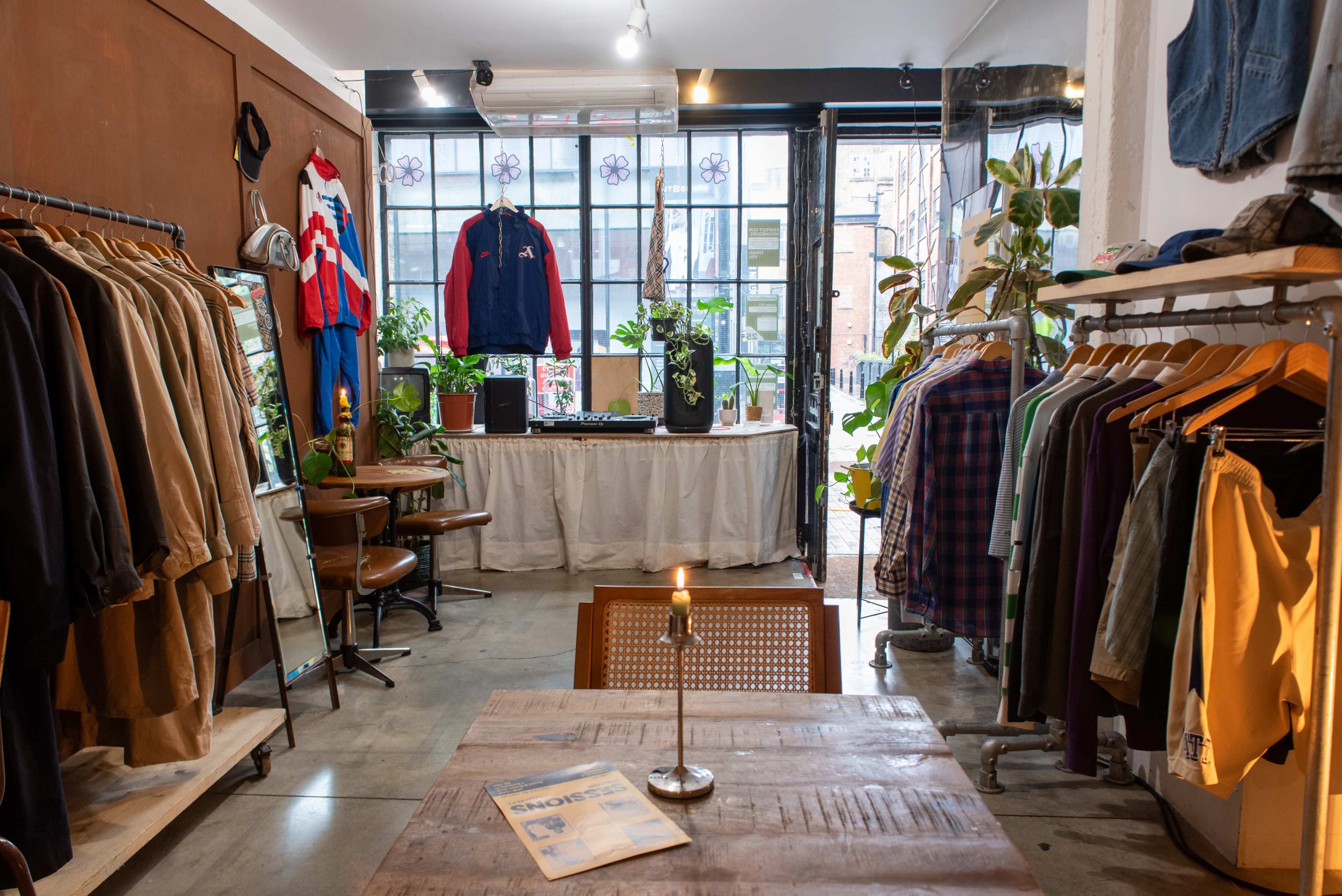 A clothing store features racks of apparel, a table in the center with a candle, and plants among the displays.
