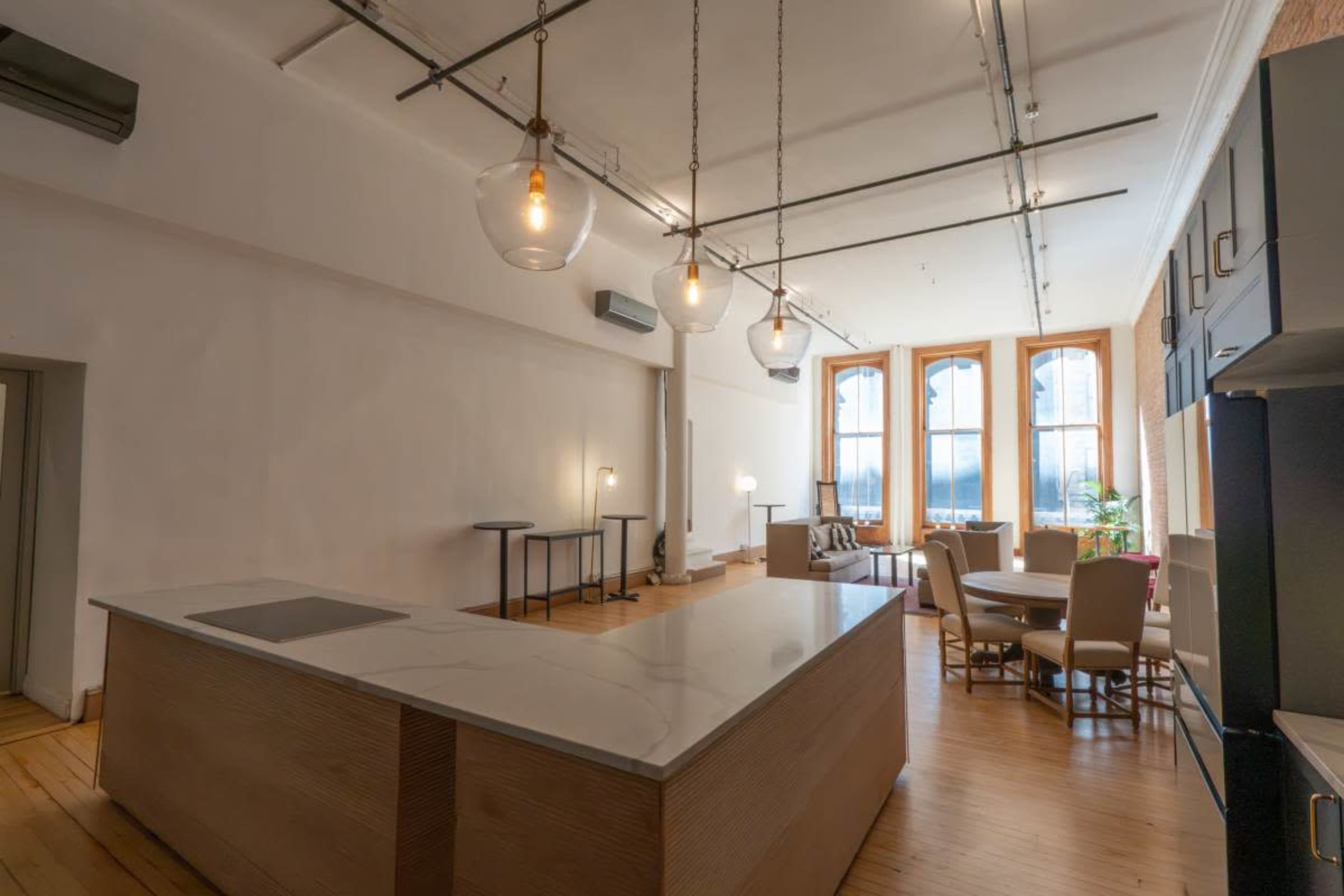 The image shows a spacious, modern kitchen area with a large marble island, pendant lighting, and large windows, leading to a dining area with seating.