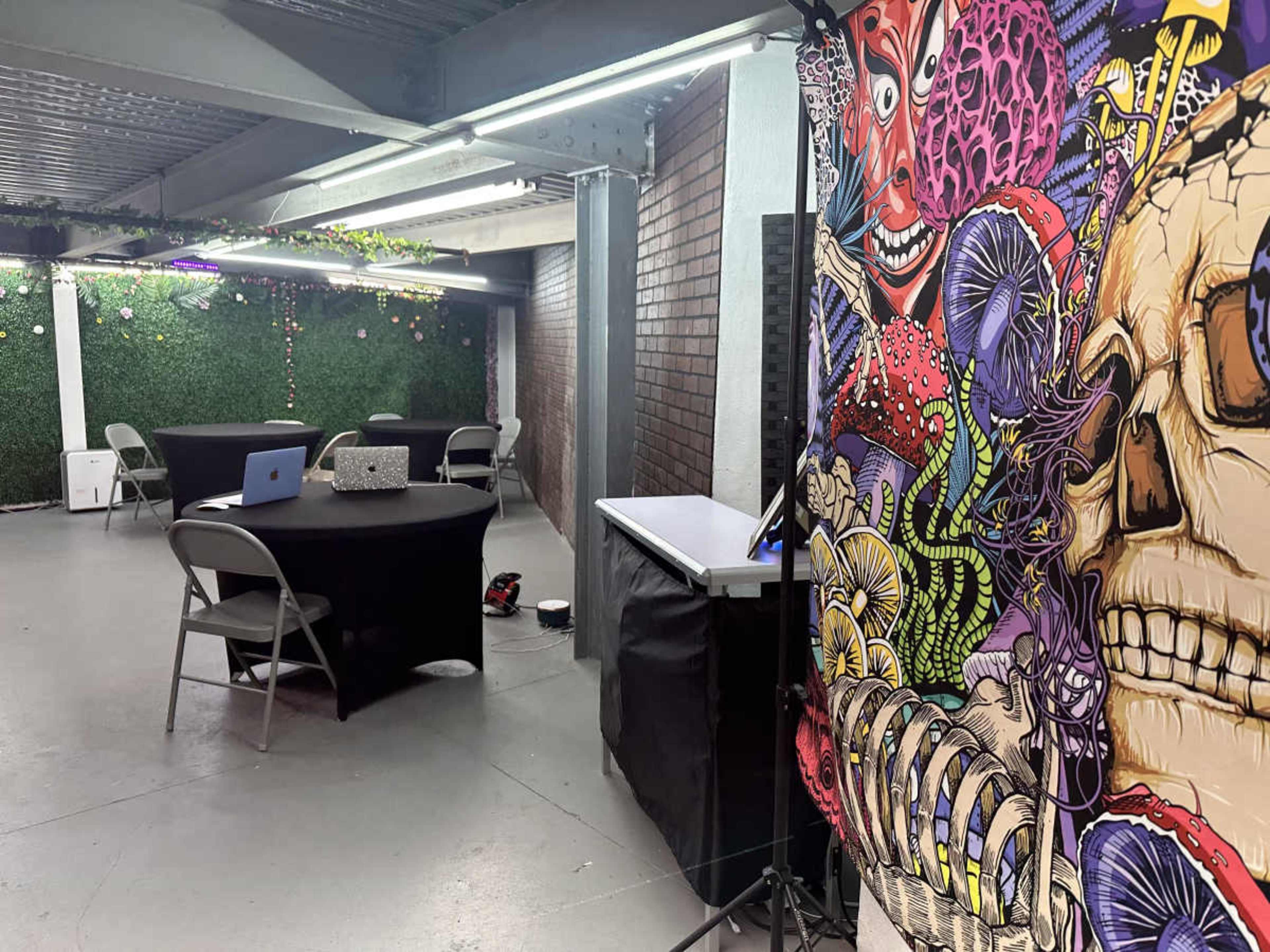 The image shows a partially decorated indoor space featuring two tables with chairs, a colorful mural on one wall, and greenery along another wall.