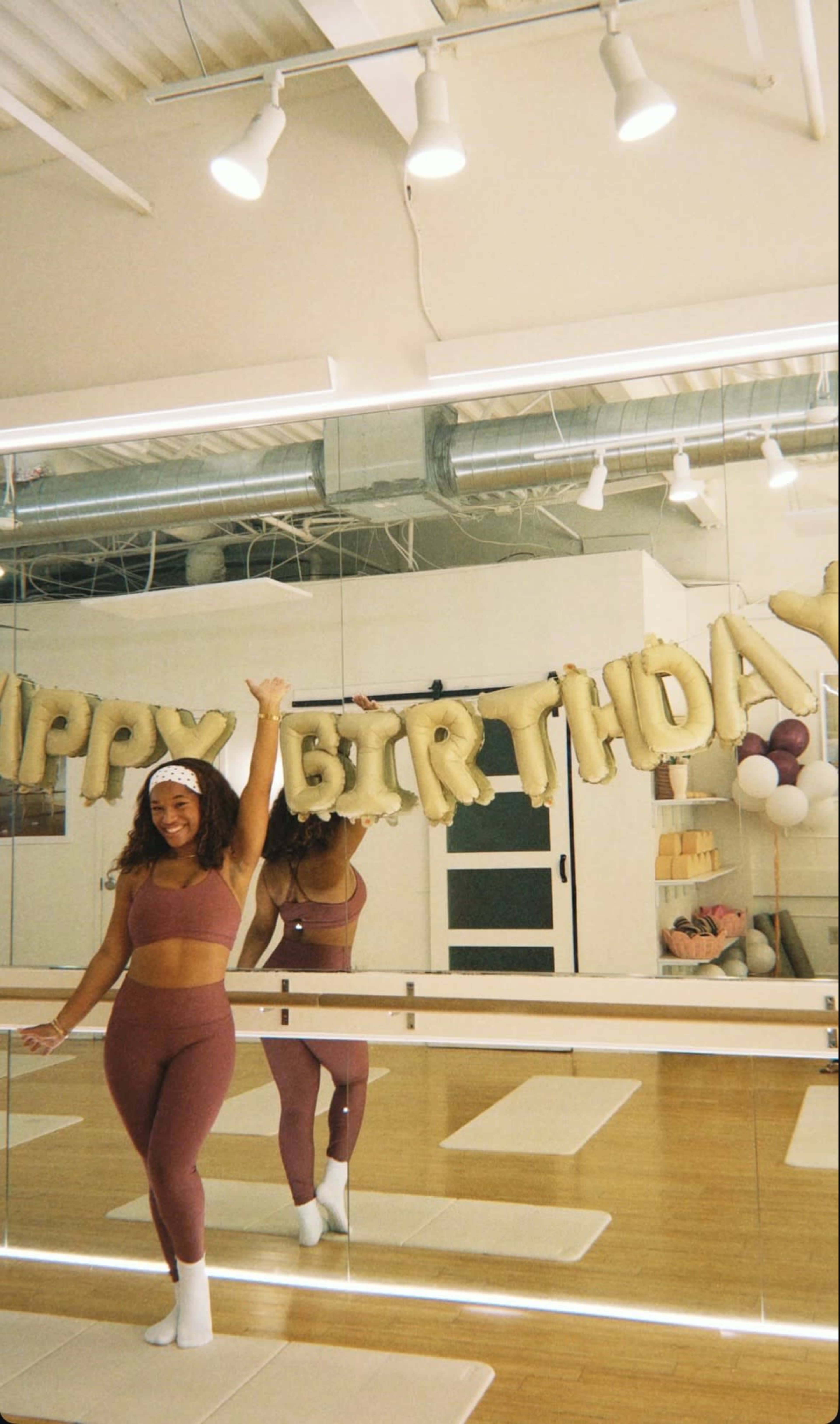 A person in fitness attire poses in front of a mirror, holding a "HAPPY BIRTHDAY" banner in a brightly lit studio space.